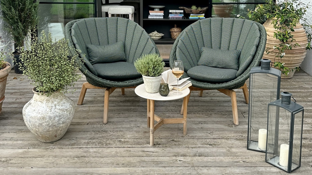 Green plant in a ceramic pot, two dark green Peacock lounge chairs with comfy seat cushions, small side tables with white ceramic table top and teak base, two black lanterns, outdoor terrace