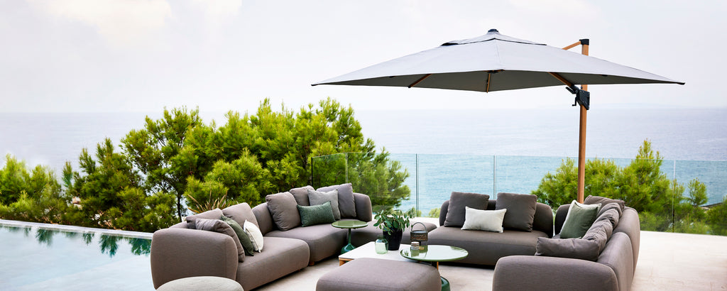 Modern seating area with gray cushions, accented by a large parasol, set against a backdrop of greenery and a serene water feature.