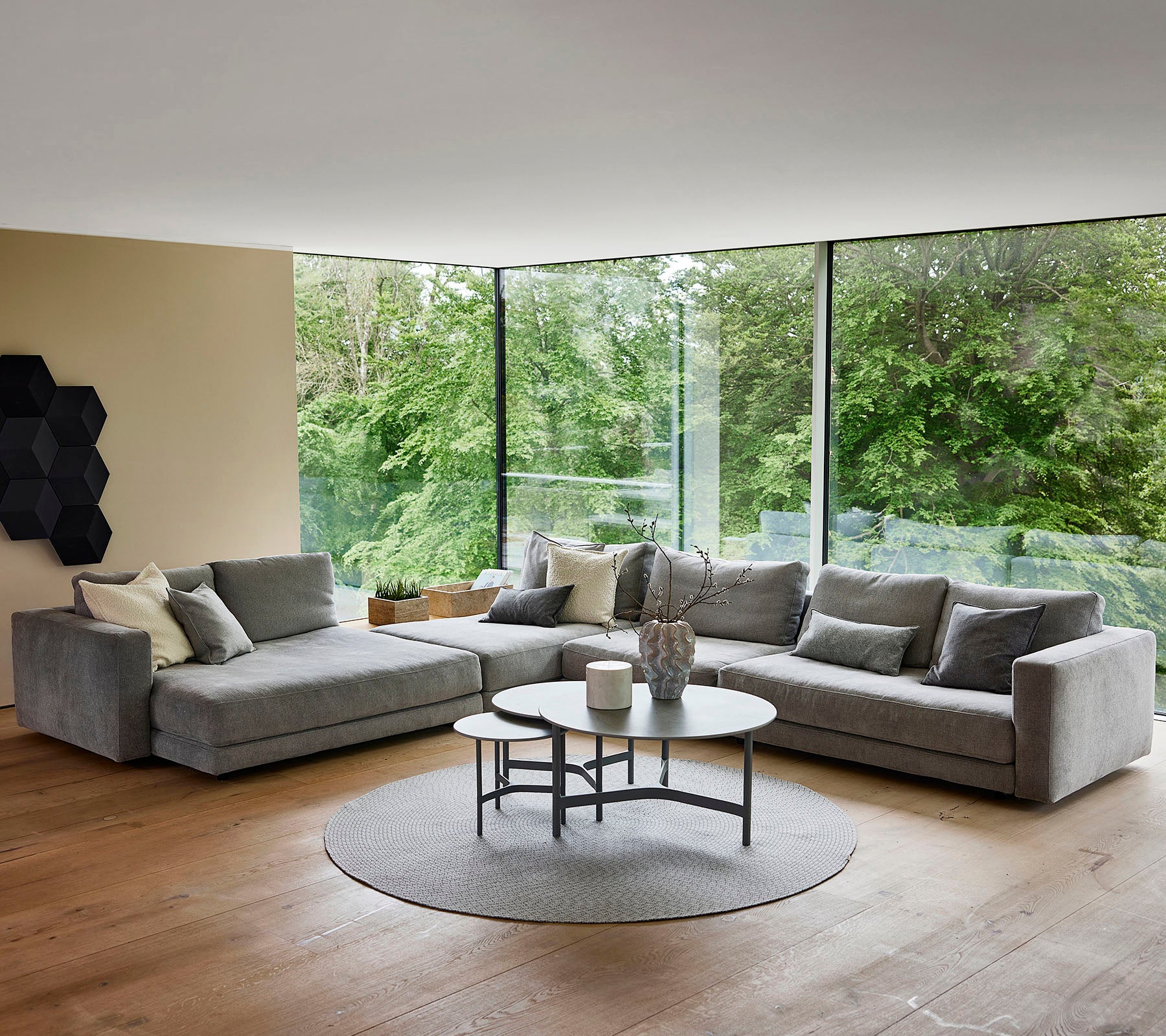 A modern living area featuring a grey sectional sofa with decorative pillows, a round coffee table, and a circular rug, surrounded by greenery.