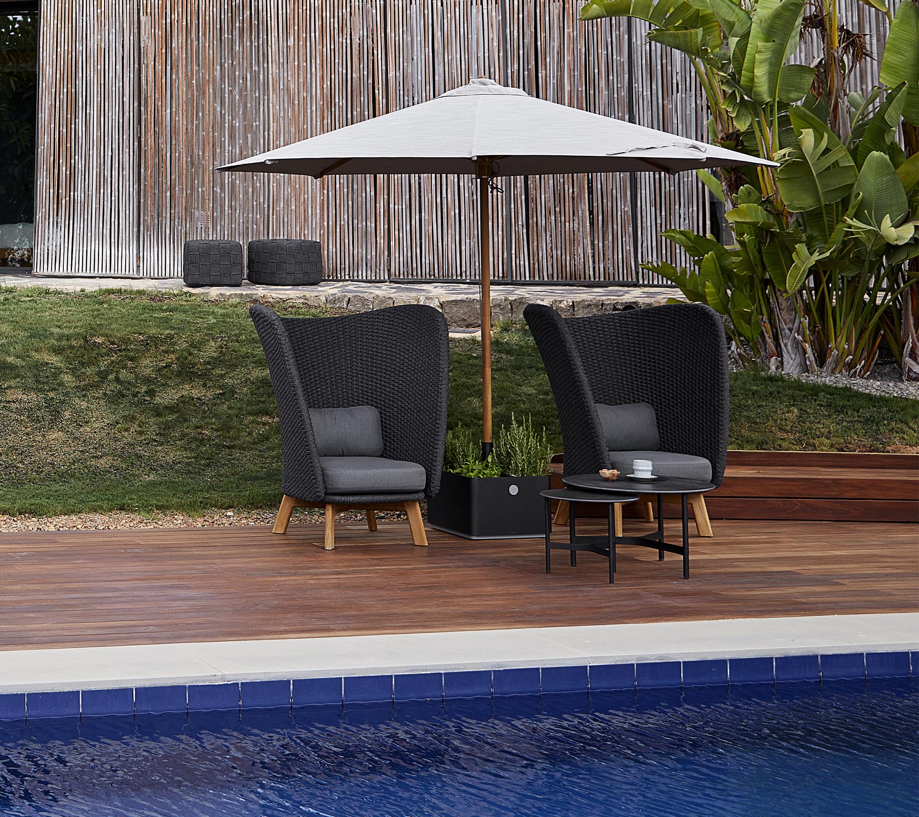 Two stylish outdoor chairs with cushions under a large umbrella, accompanied by a small table, alongside a swimming pool.