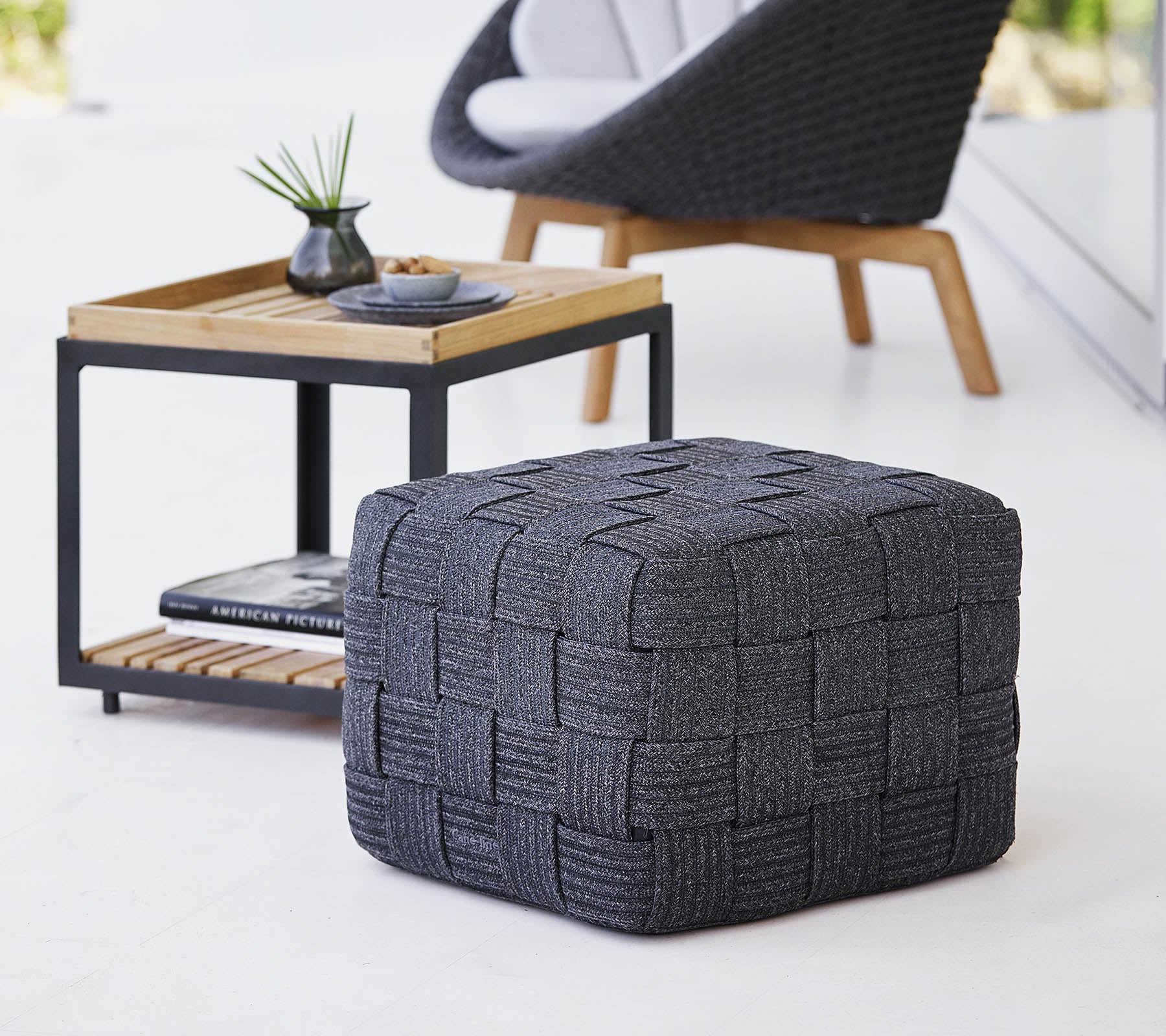 A textured gray woven pouf positioned near a wooden side table with a minimalistic design, featuring small decor items.