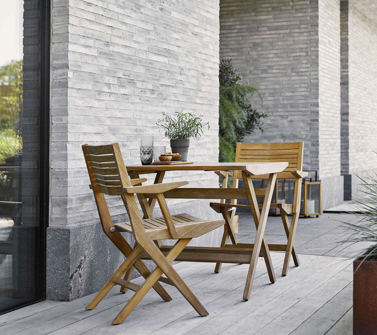 A wooden folding table with two matching chairs, adorned with potted plants, against a textured stone wall.