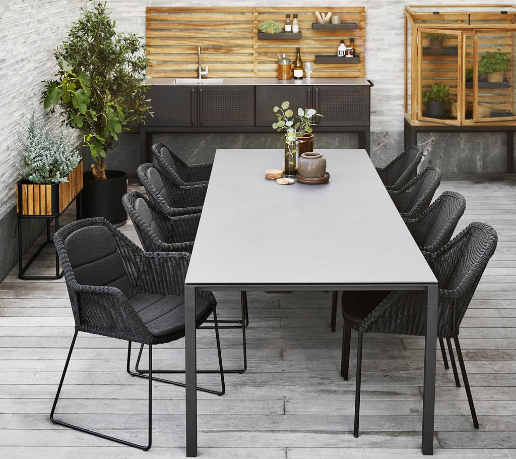 A modern dining setup featuring a long table surrounded by sleek black chairs, complemented by decorative plants and a wooden backdrop.