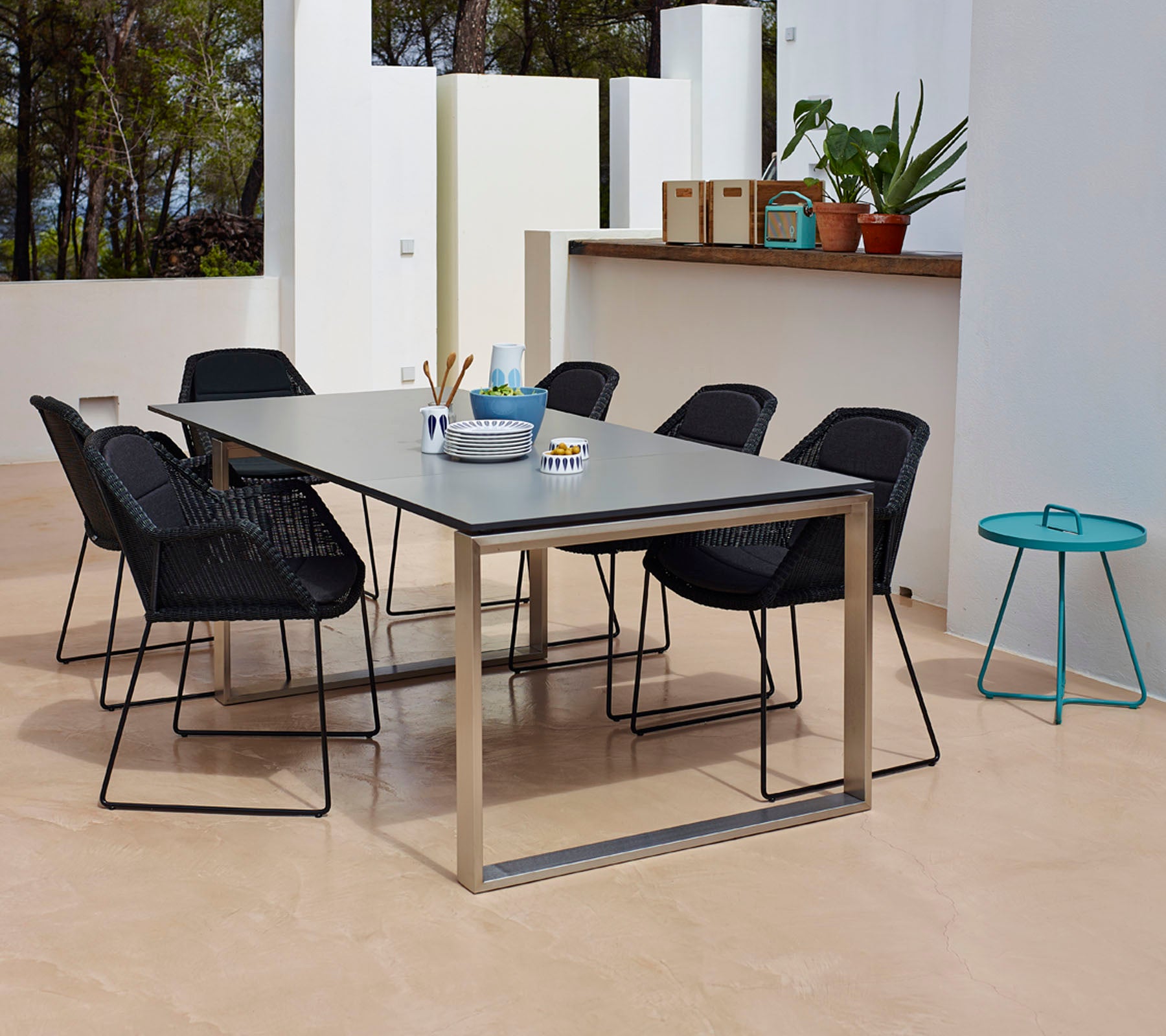 A modern dining setup featuring a rectangular table with a sleek surface, surrounded by six black woven chairs and a small teal side table.