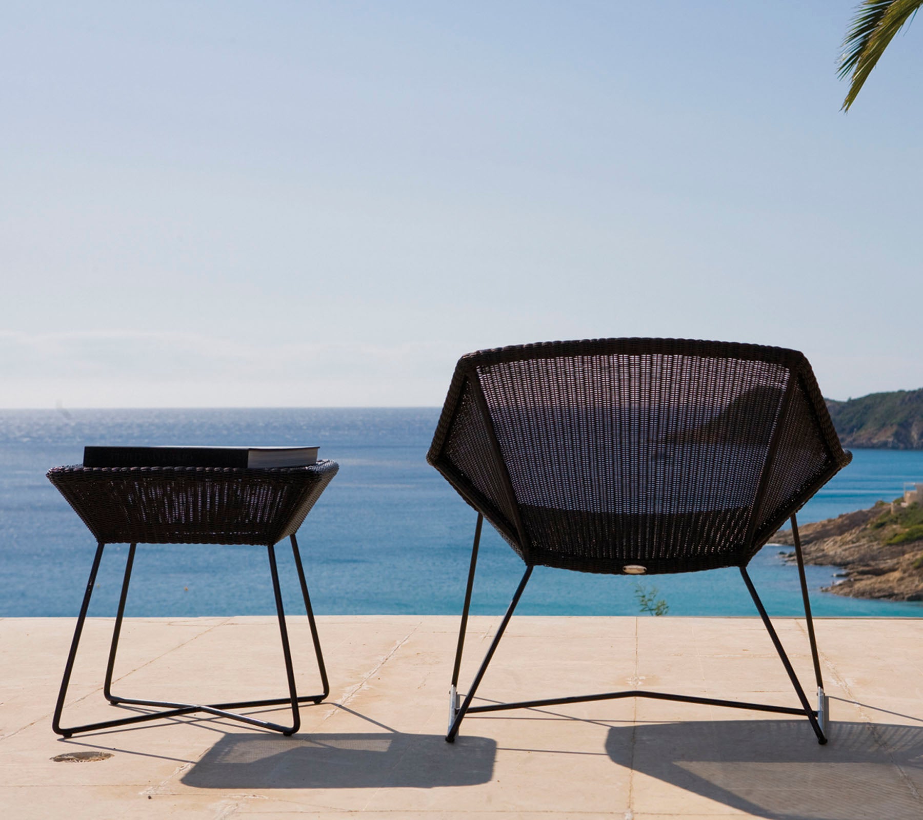 Two modern, black outdoor chairs with a geometric design are positioned beside a table, overlooking a calm sea under a clear sky.