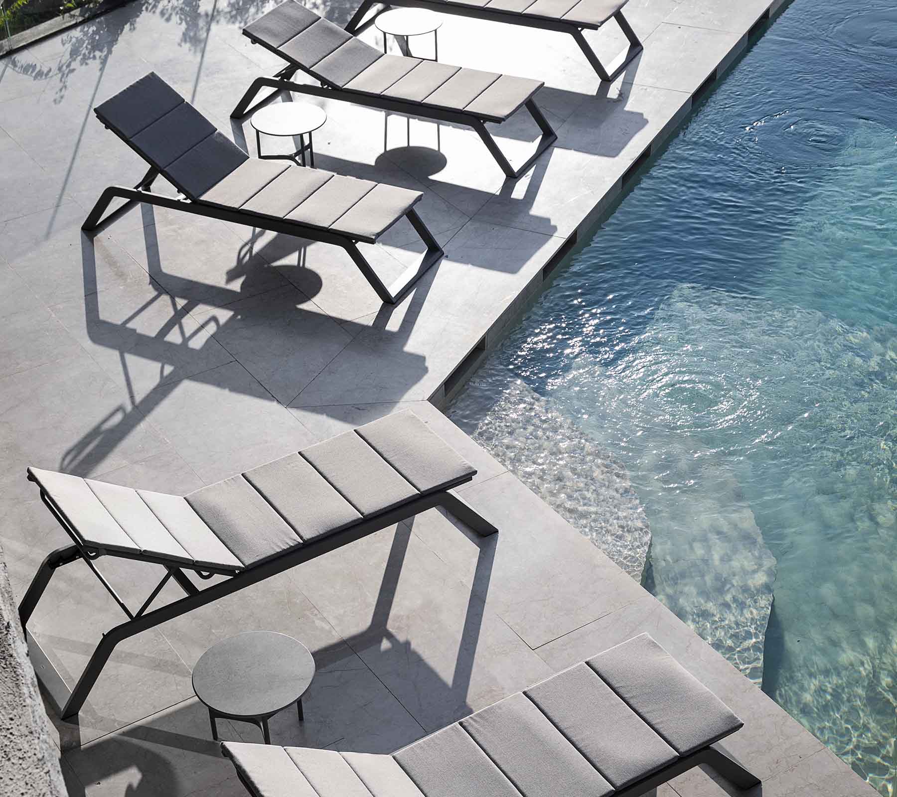 A row of modern lounge chairs with a small side table beside a swimming pool, casting shadows on the pavement.