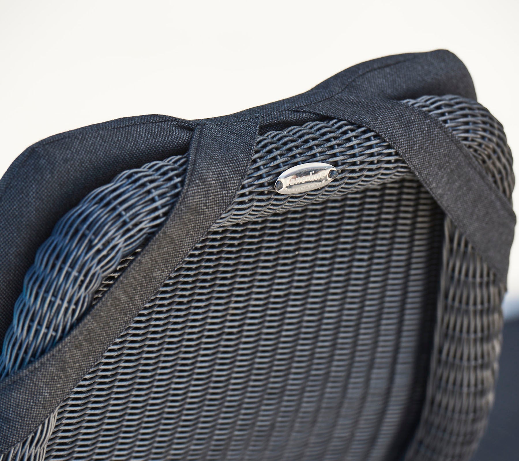 Close-up of a modern chair's back, featuring textured wicker material and a sleek, metallic logo detail.