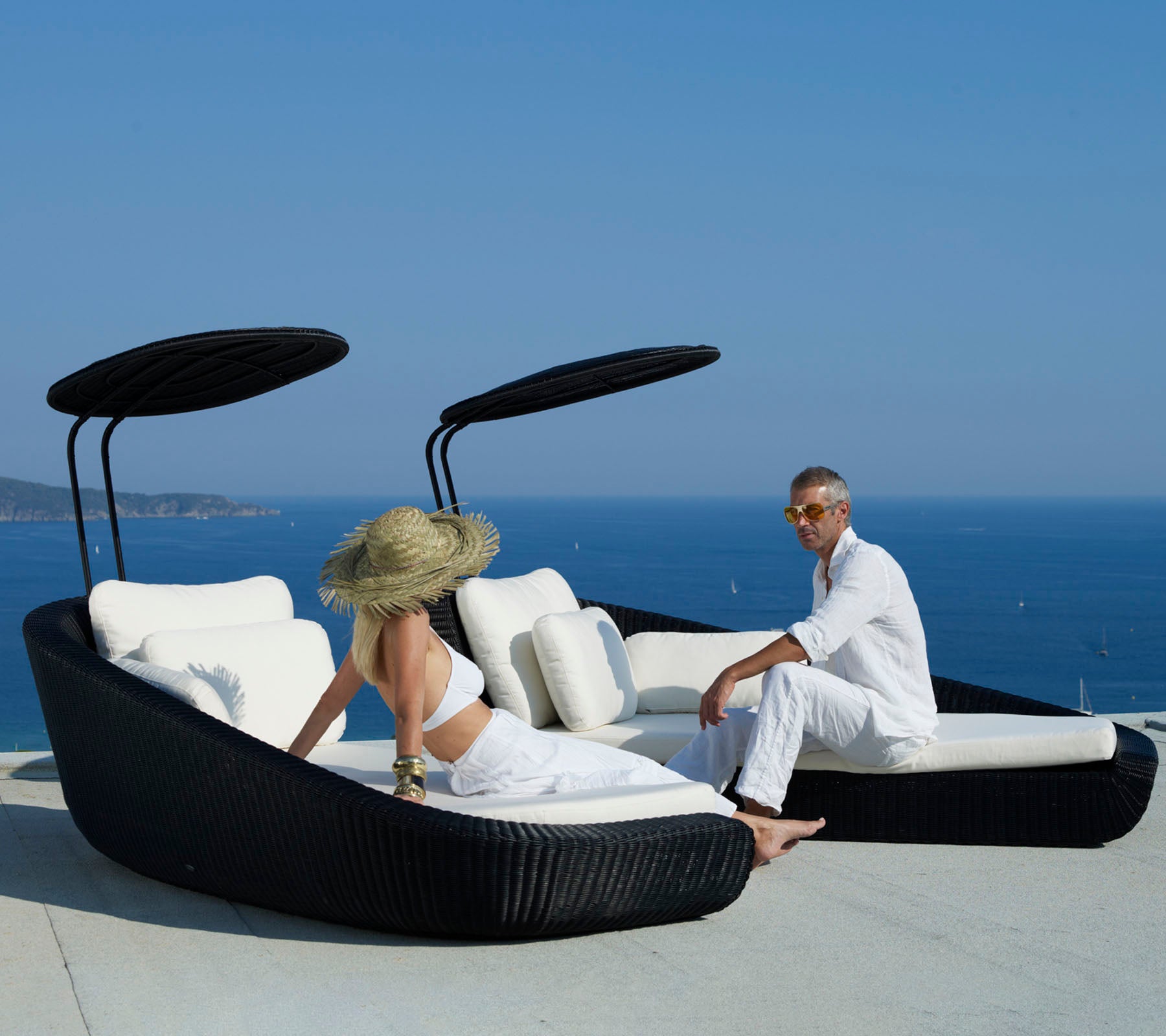 Two people relax on a stylish, curved sunbed with a scenic ocean view, featuring sleek black design and plush white cushions.