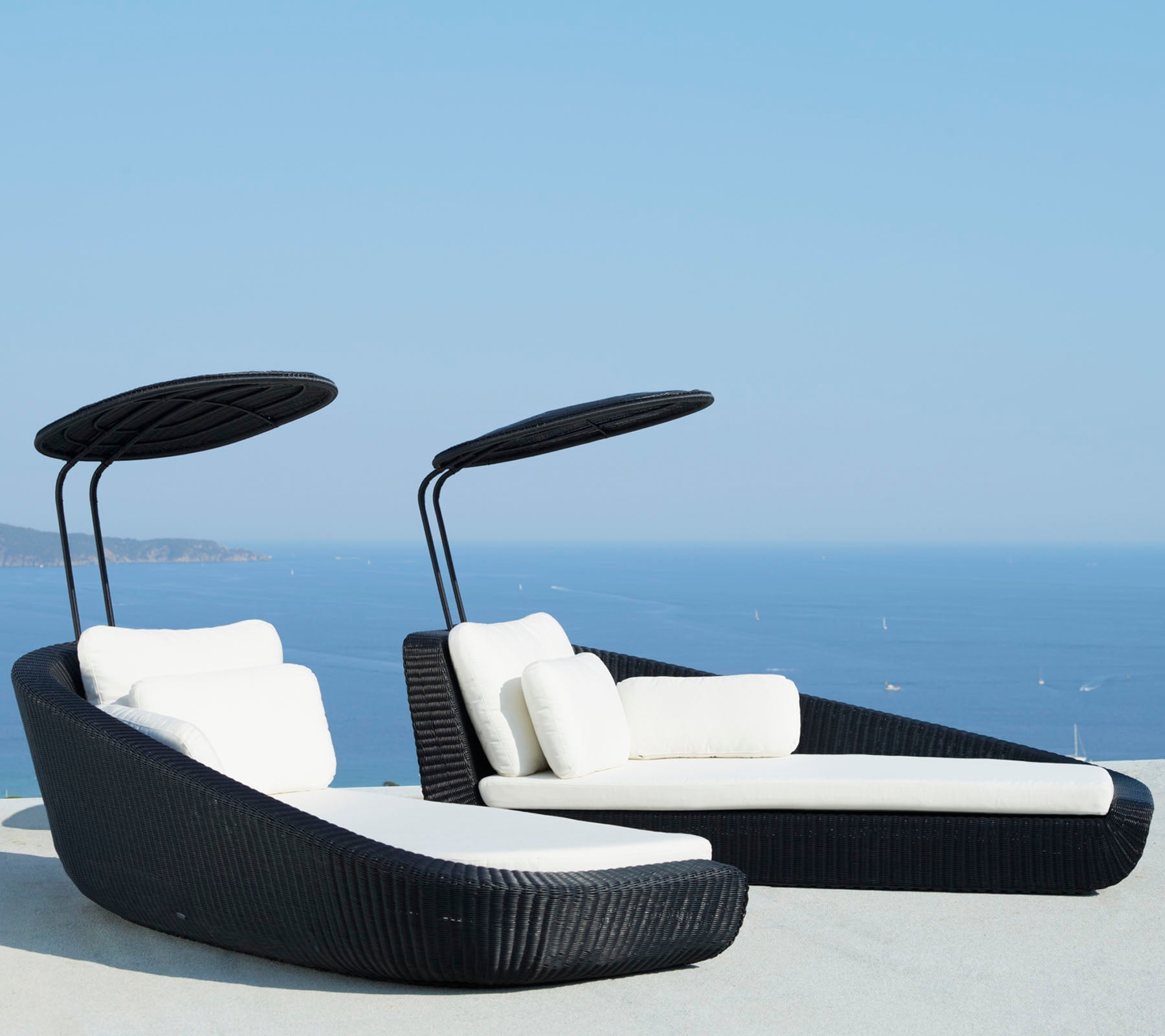 Two stylish lounge chairs with white cushions and curved canopies, set against a scenic blue ocean backdrop.