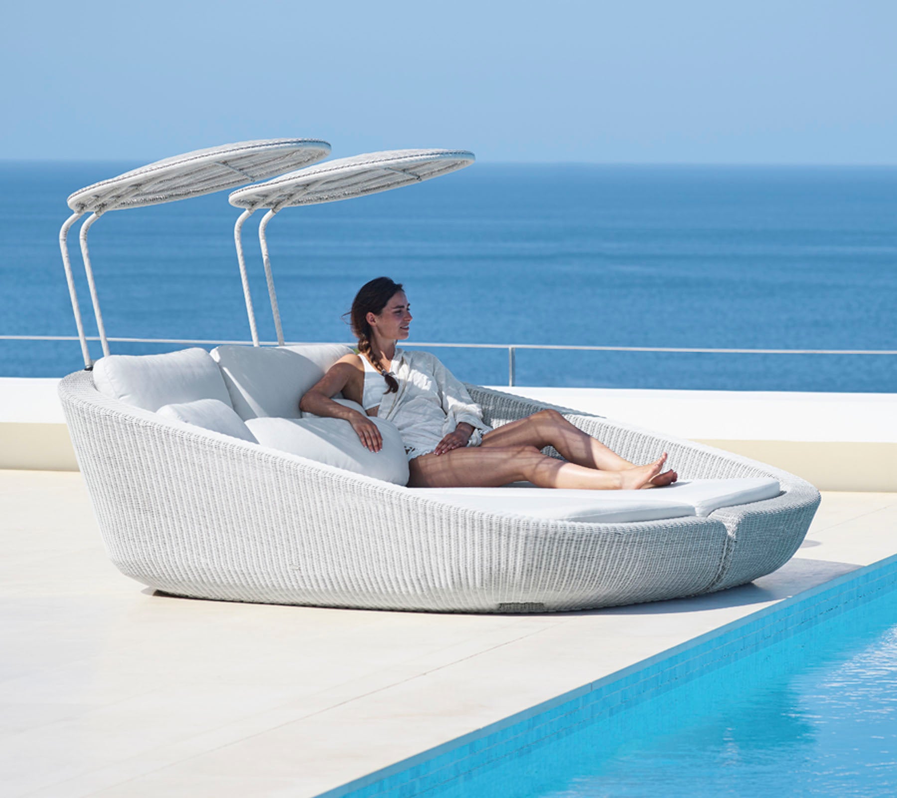 A woman relaxes on a stylish, modern lounge chair with adjustable canopies beside a tranquil body of water.