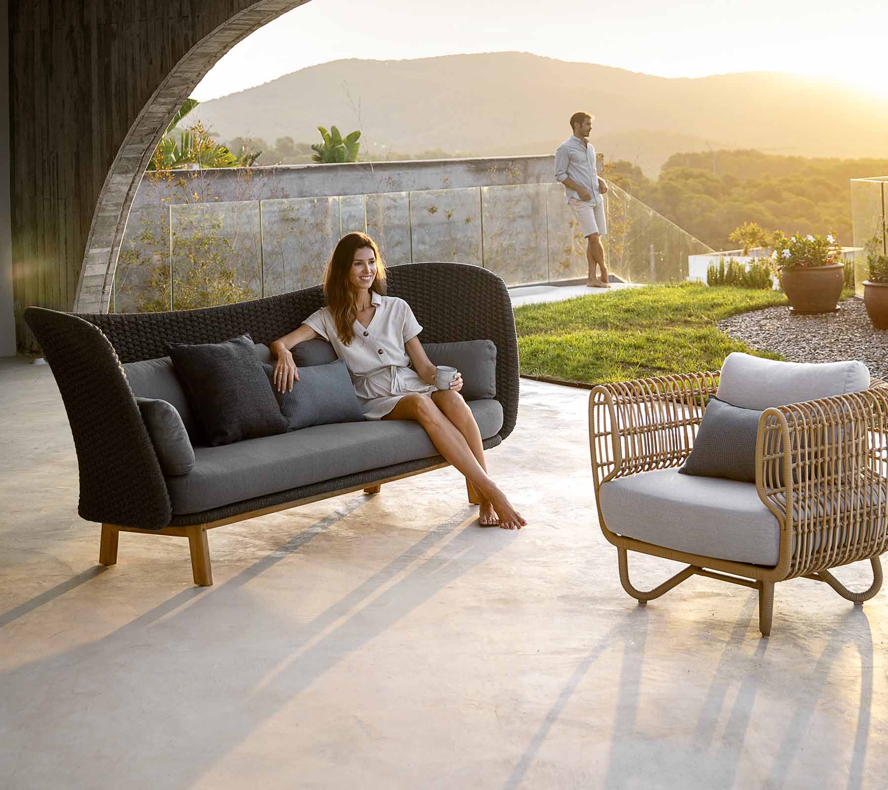 A stylish seating arrangement featuring a woman relaxing on a modern sofa with diverse cushions, and a chair nearby, set against a scenic backdrop.