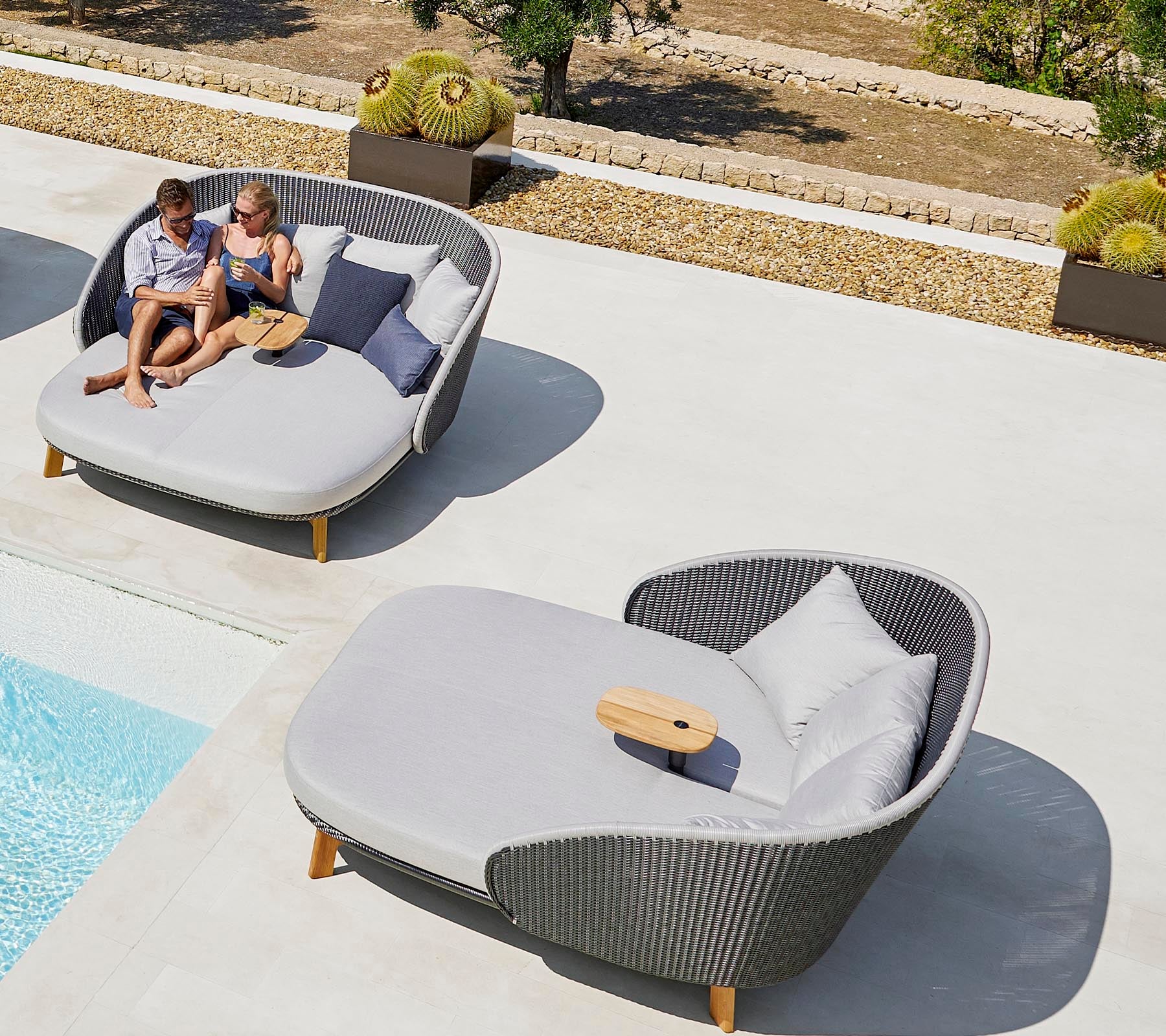Two people relax on stylish outdoor loungers near a swimming pool, enjoying leisure time with cushions and a small table.