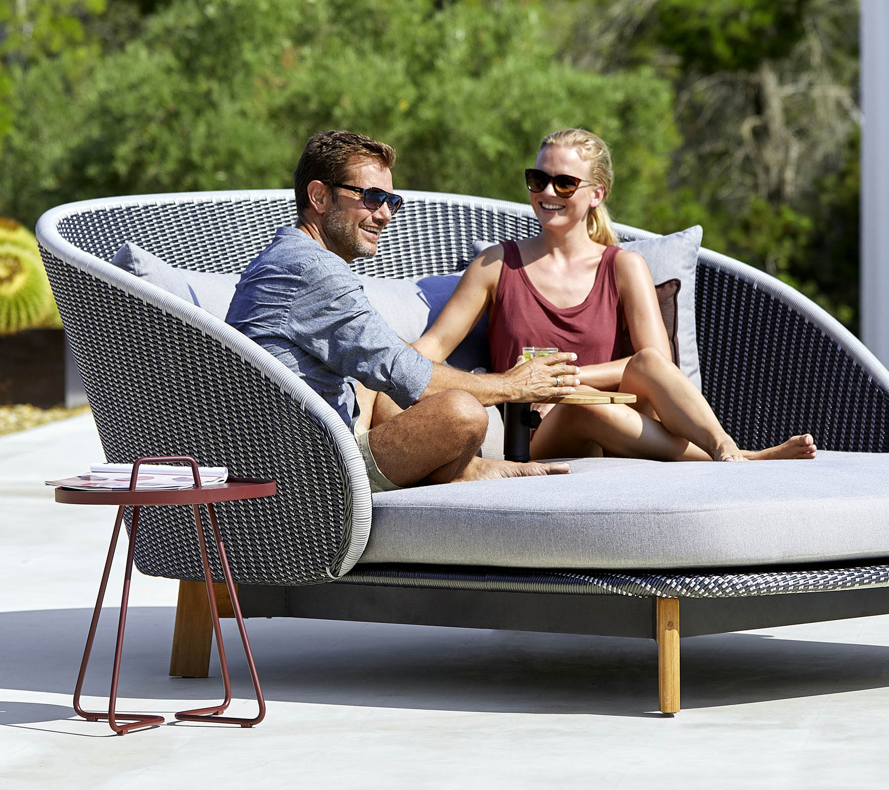 A couple enjoys a cozy moment together on a stylish, curved outdoor sofa with a side table nearby.