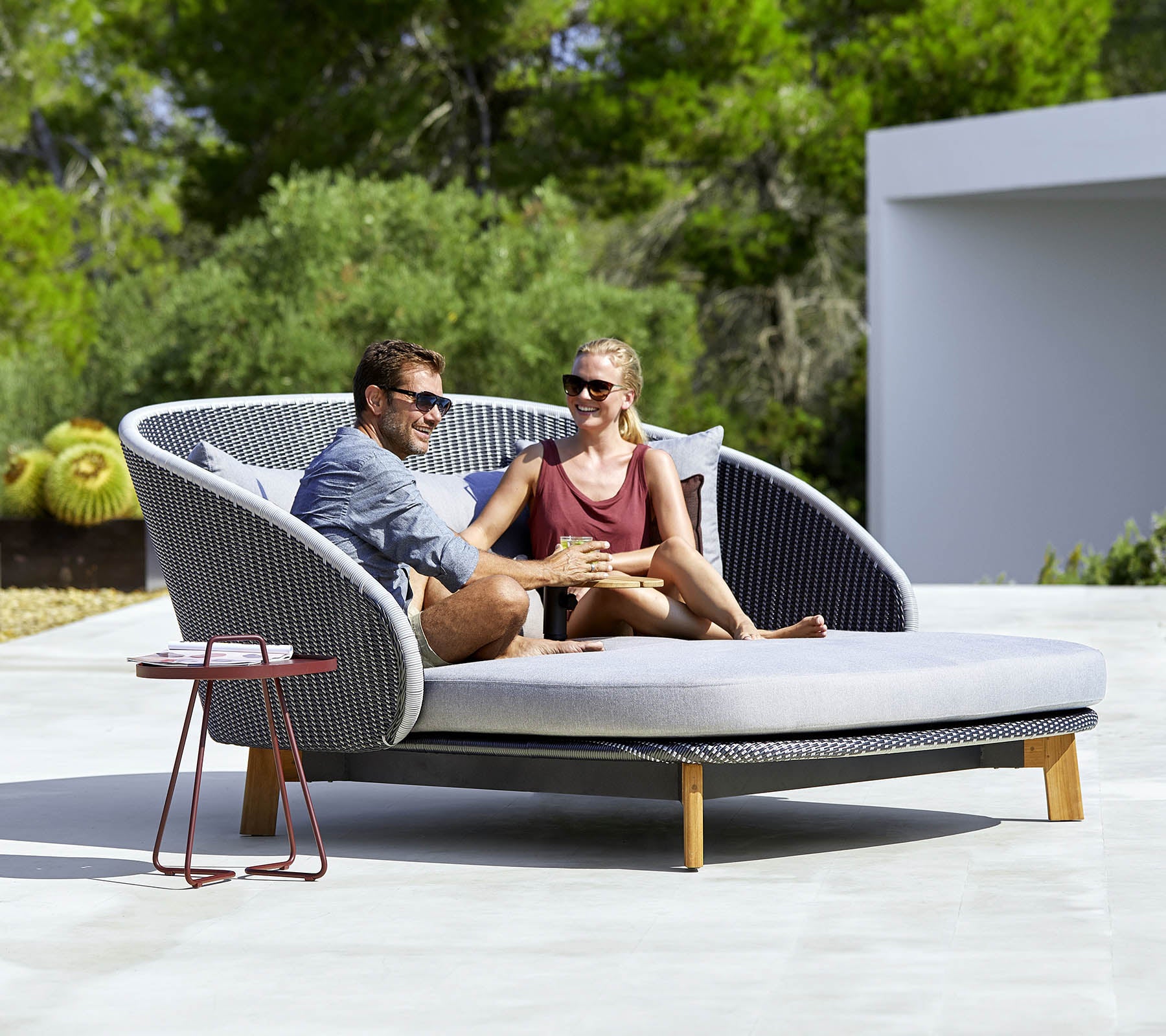 A stylish, curved outdoor sofa with two people relaxing together, surrounded by greenery and a small side table nearby.