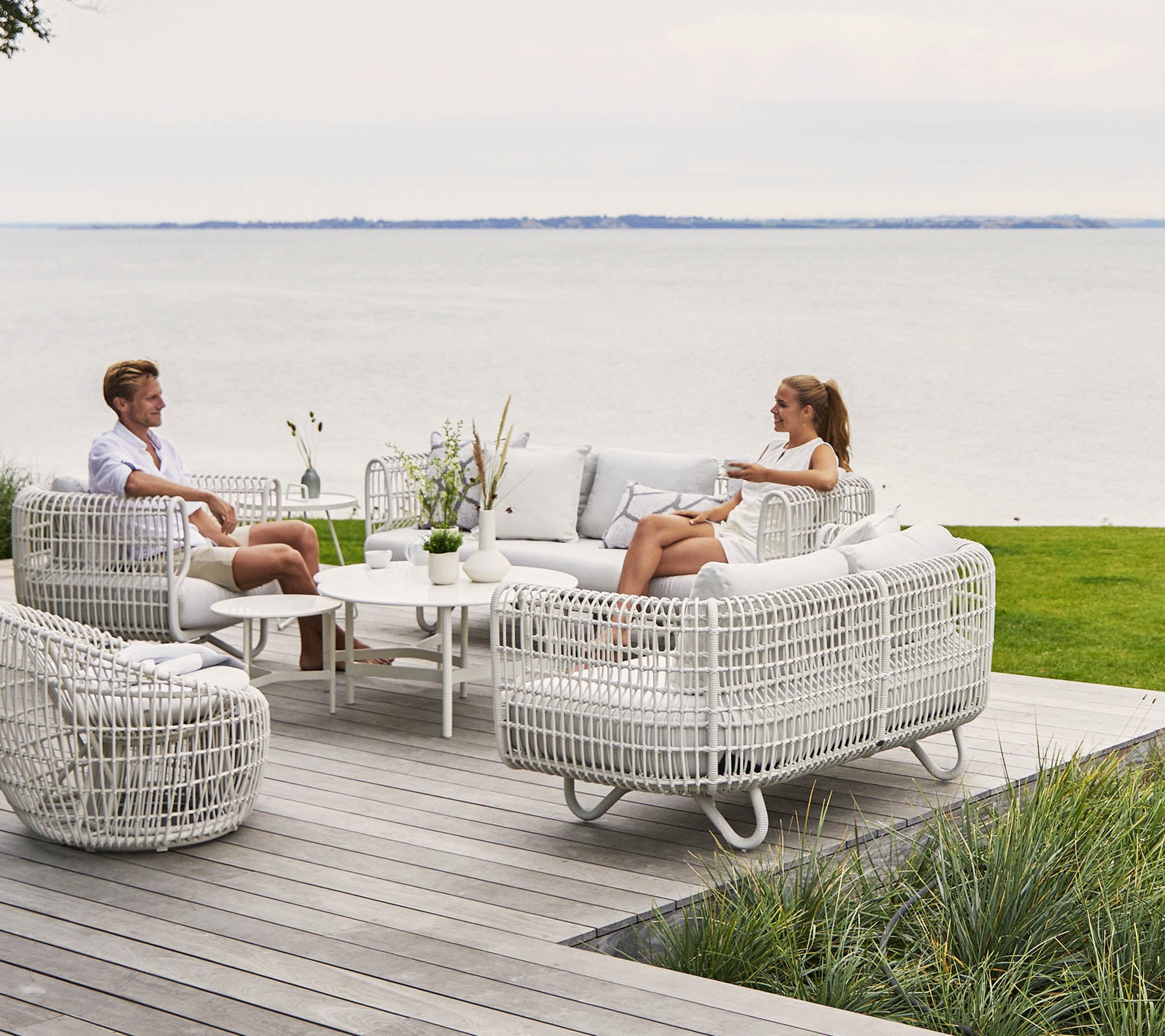 Two people relax on a stylish, comfortable seating area with white wicker furniture and a coffee table, overlooking a scenic water view.