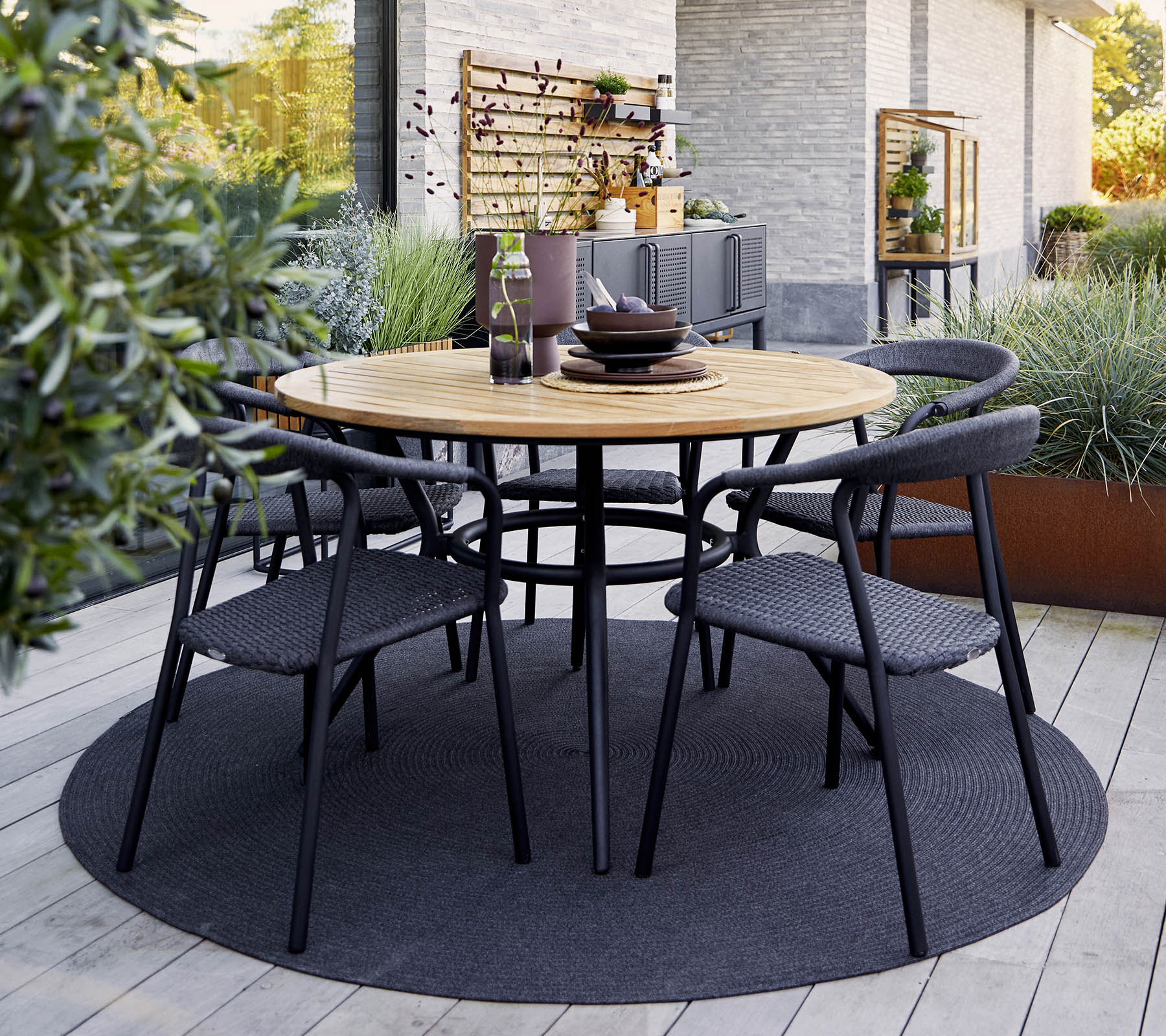 Round wooden table with black chairs arranged around it, set for dining, surrounded by greenery and a textured rug.