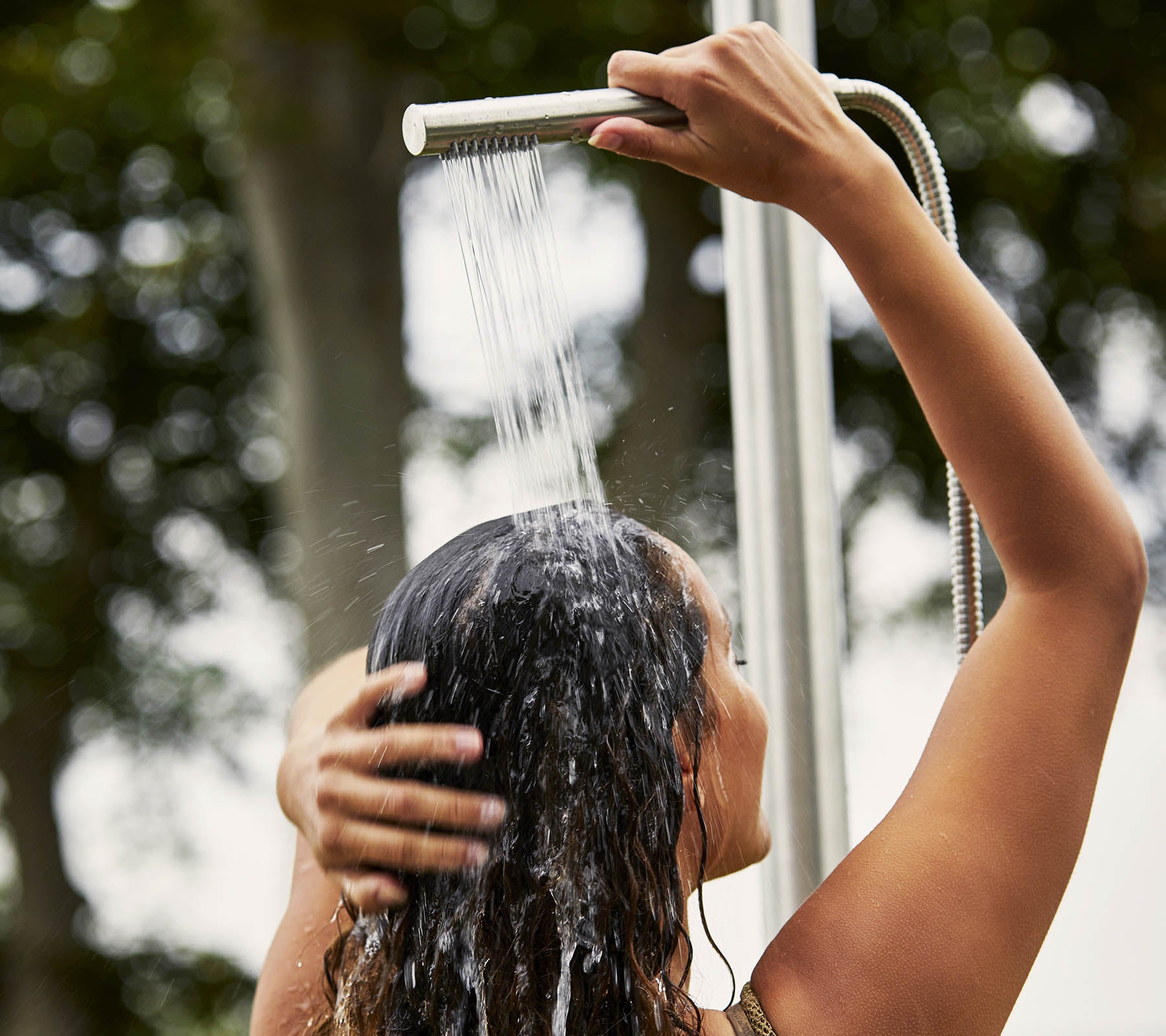 A person washing their hair under a shower, with water cascading down and surrounded by a natural setting.