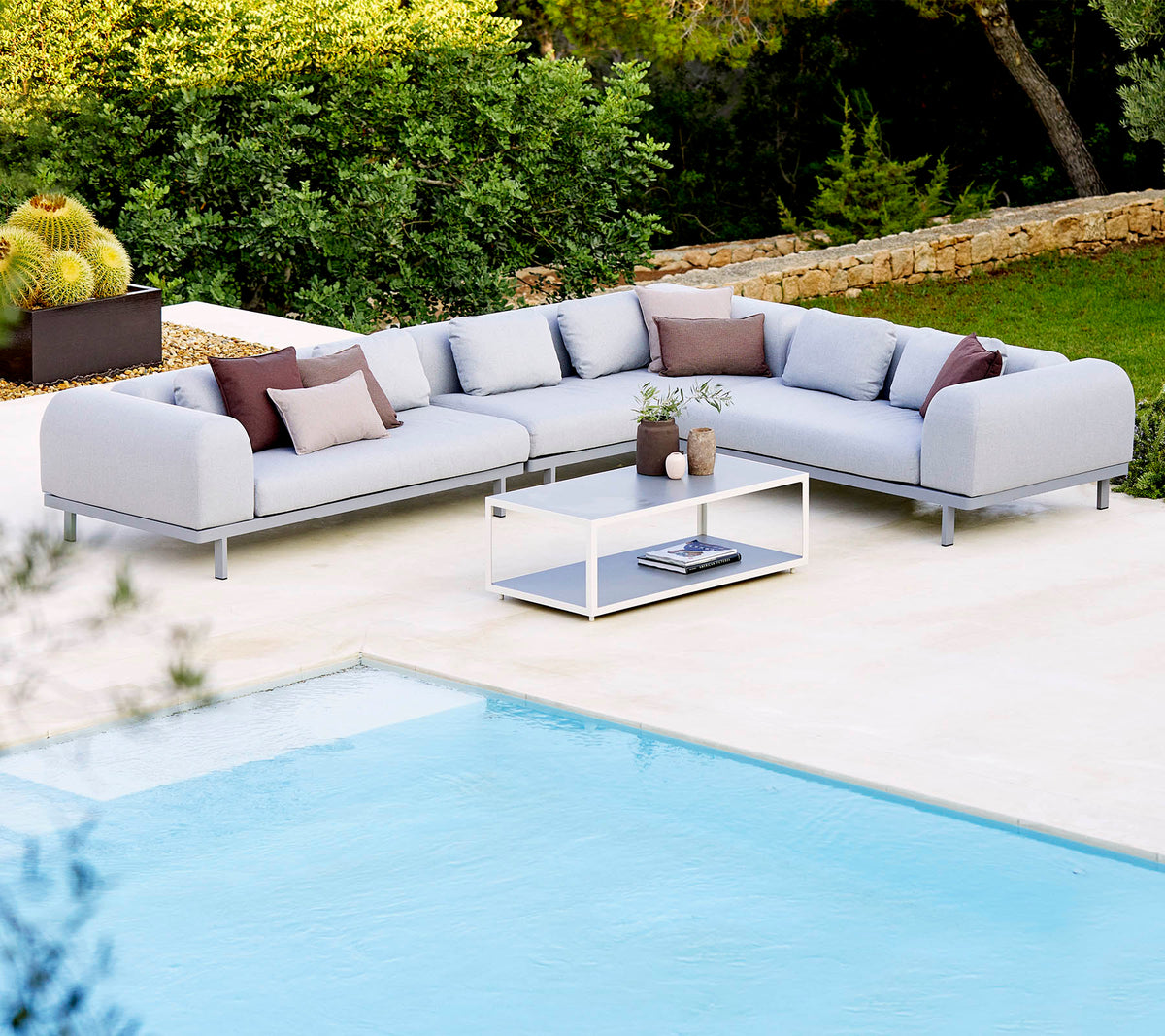 Light grey sectional sofa with decorative cushions and a glass coffee table, positioned next to a swimming pool and lush greenery.