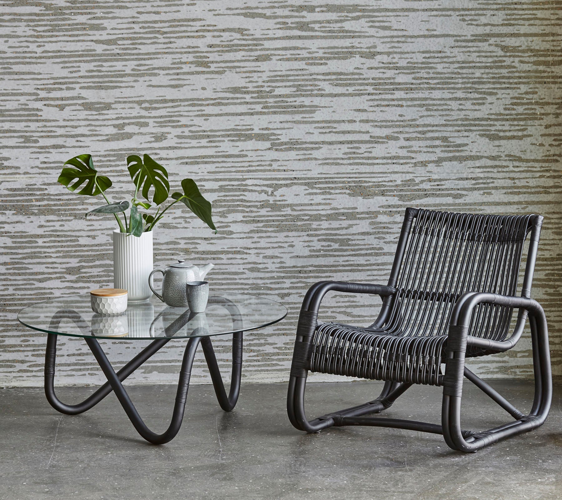 A stylish chair beside a glass coffee table, featuring a vase with greenery and a small cup of tea, set against a textured wall.