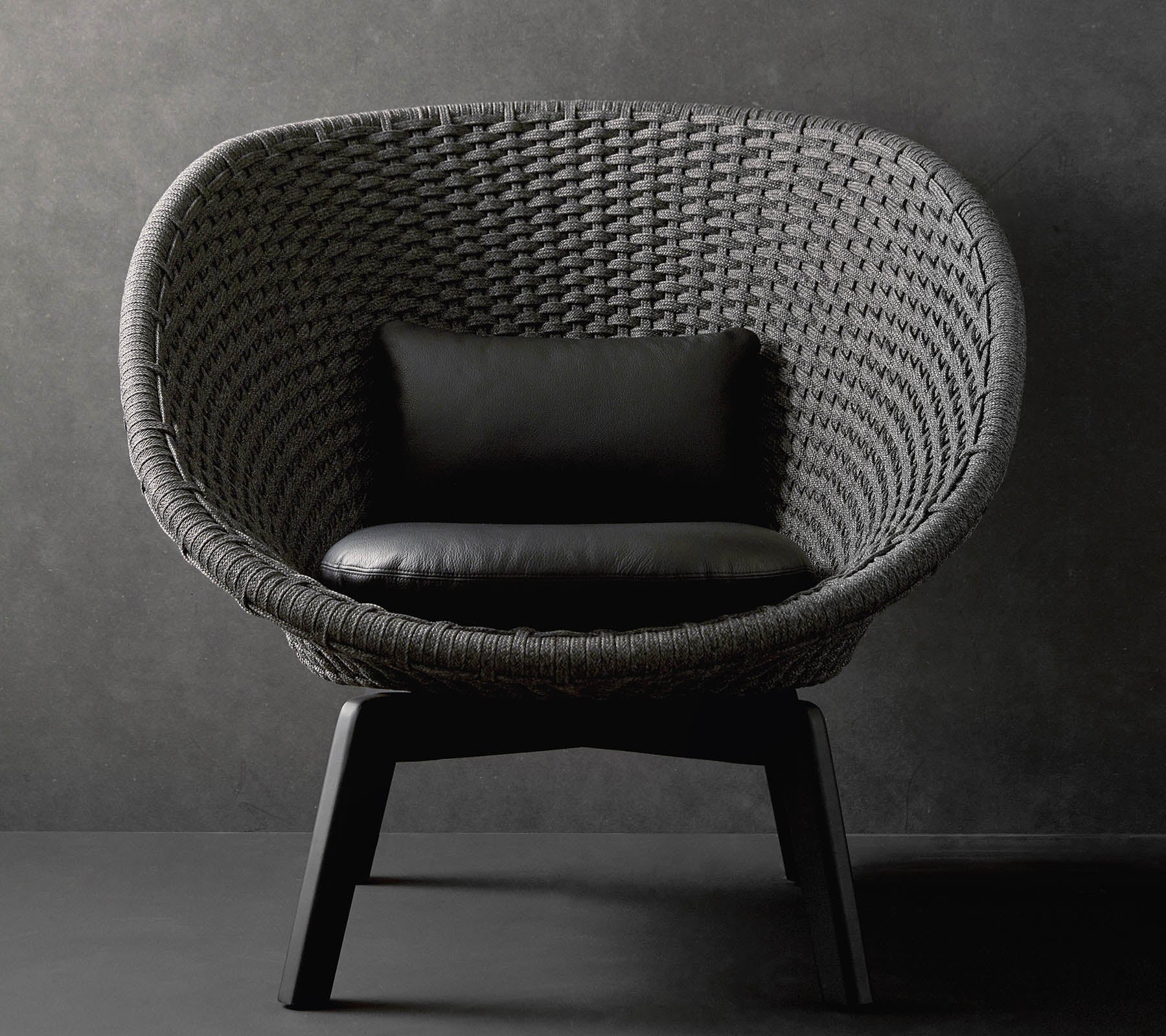 A stylish round chair with a textured woven design and a black cushion, resting on sleek wooden legs against a dark background.