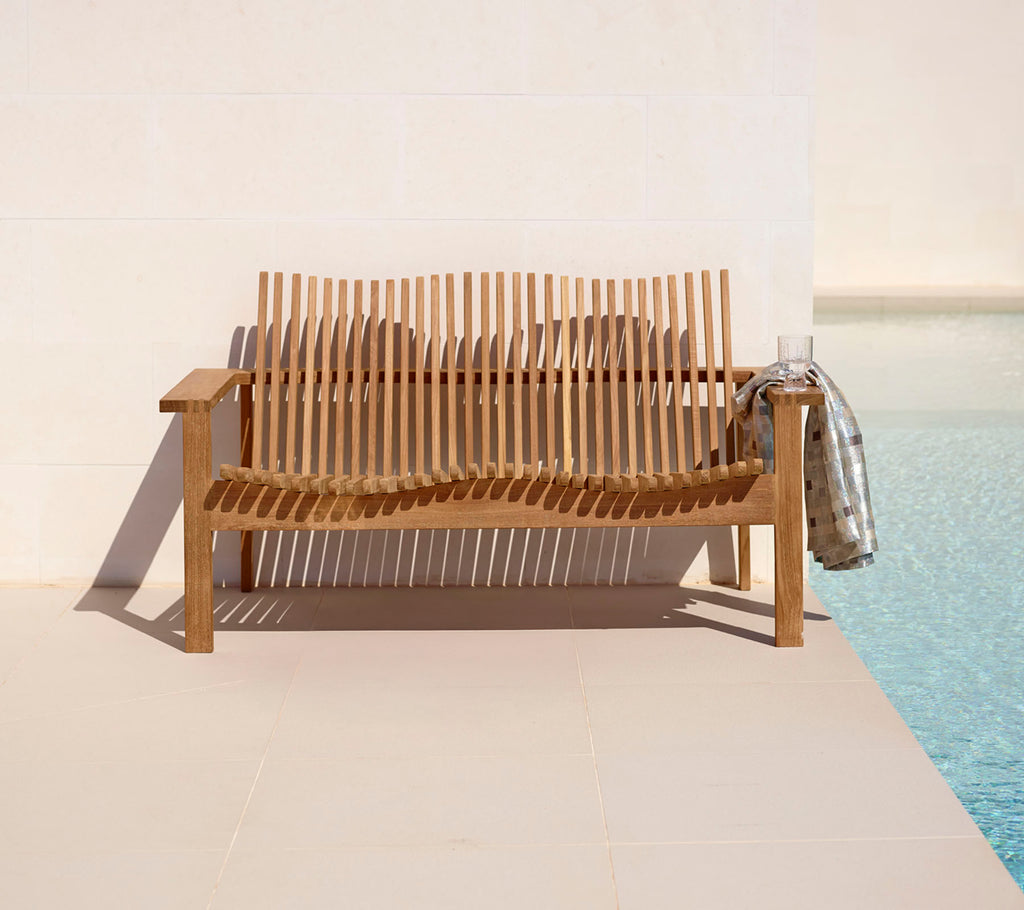 Stylish wooden 2-seater sofa with slatted design, complemented by a cozy throw, beside a tranquil pool. Perfect for relaxation.