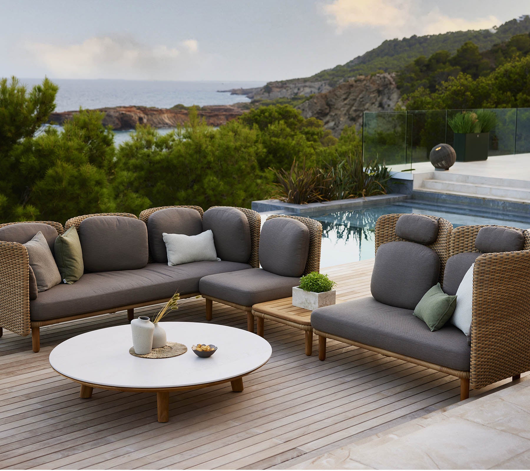 Stylish lounge seating with gray cushions and decorative pillows, accompanied by a round table, set against a scenic backdrop.