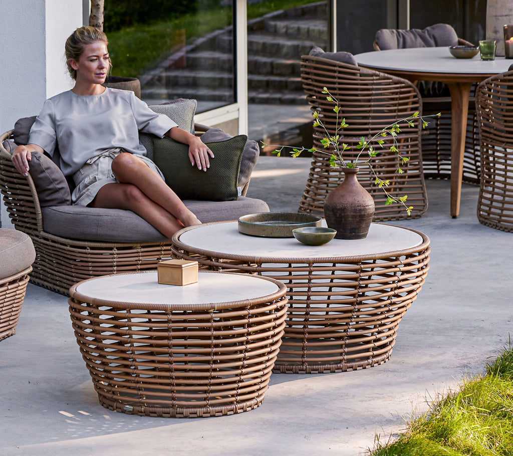 A seated person relaxes beside medium and large woven basket coffee tables, featuring a blend of natural materials and minimalist design.
