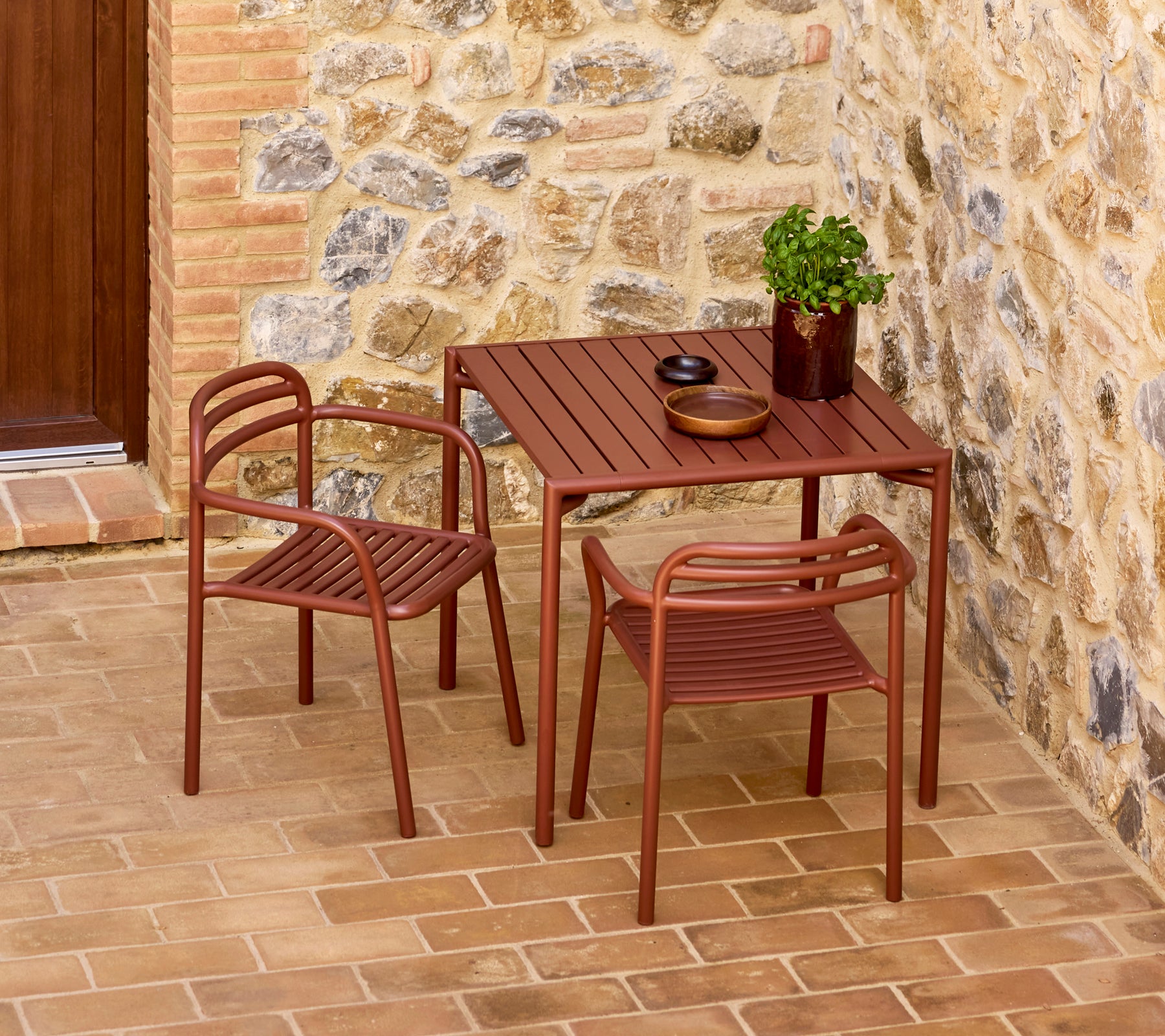 Stylish desert red Bliss table with two matching chairs, accompanied by a decorative plant and simple tableware against a stone backdrop.