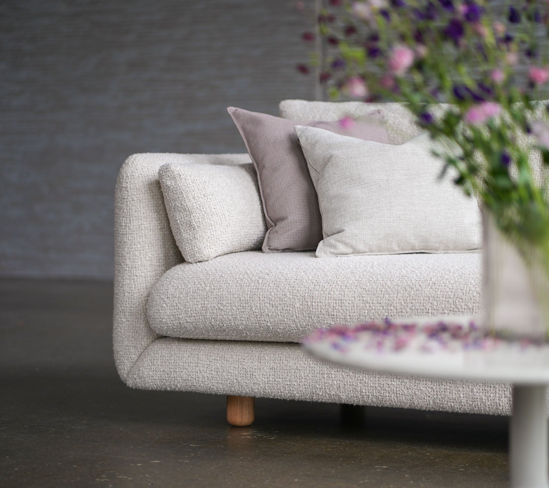 Soft sofa with sand-colored upholstery, decorative pillows, teak legs, and a vase of flowers, showcasing Cane-line flair and elegance.