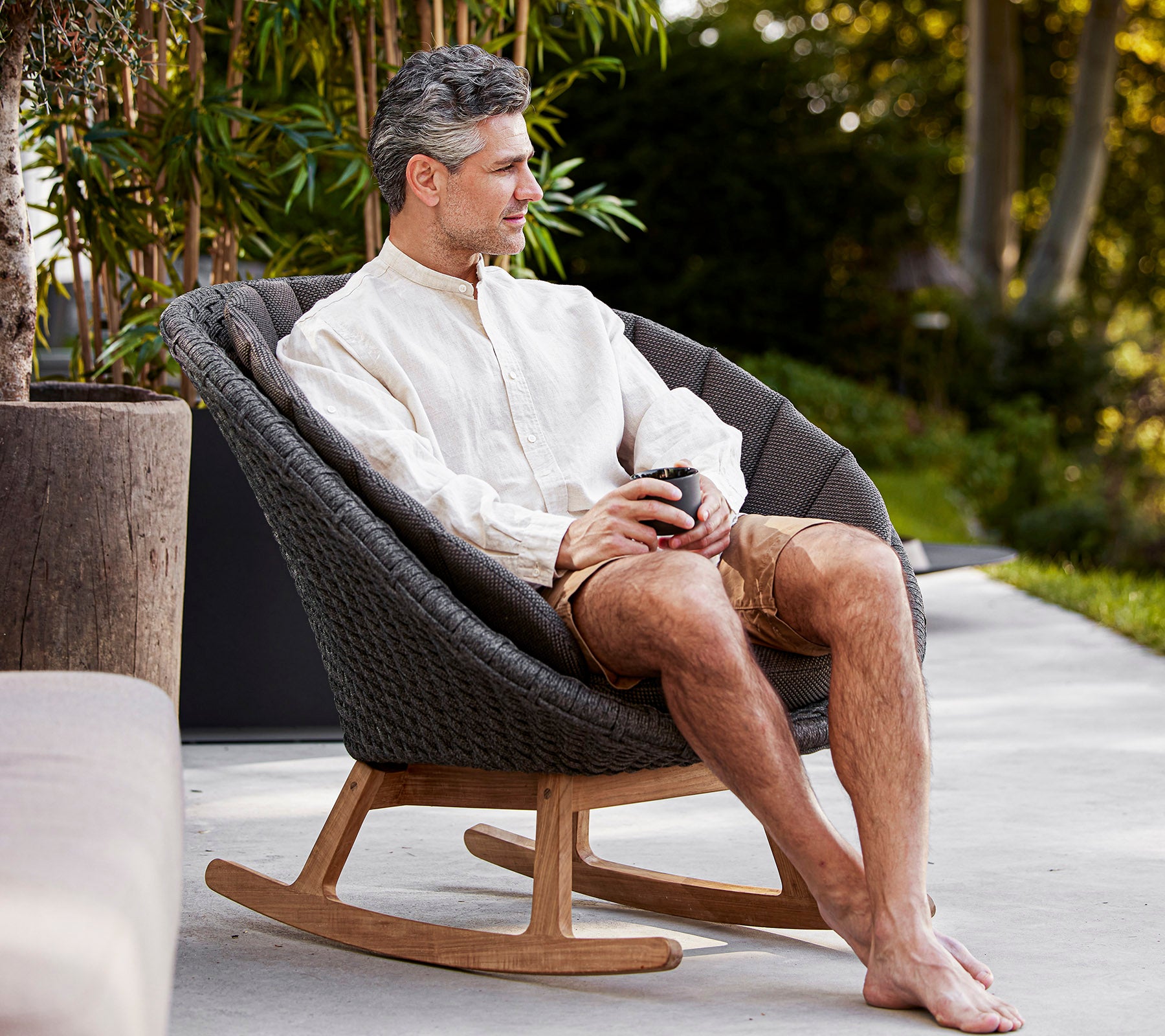A man sits comfortably in a stylish rocking chair, enjoying a drink in a serene setting, dressed casually in a light shirt and shorts.
