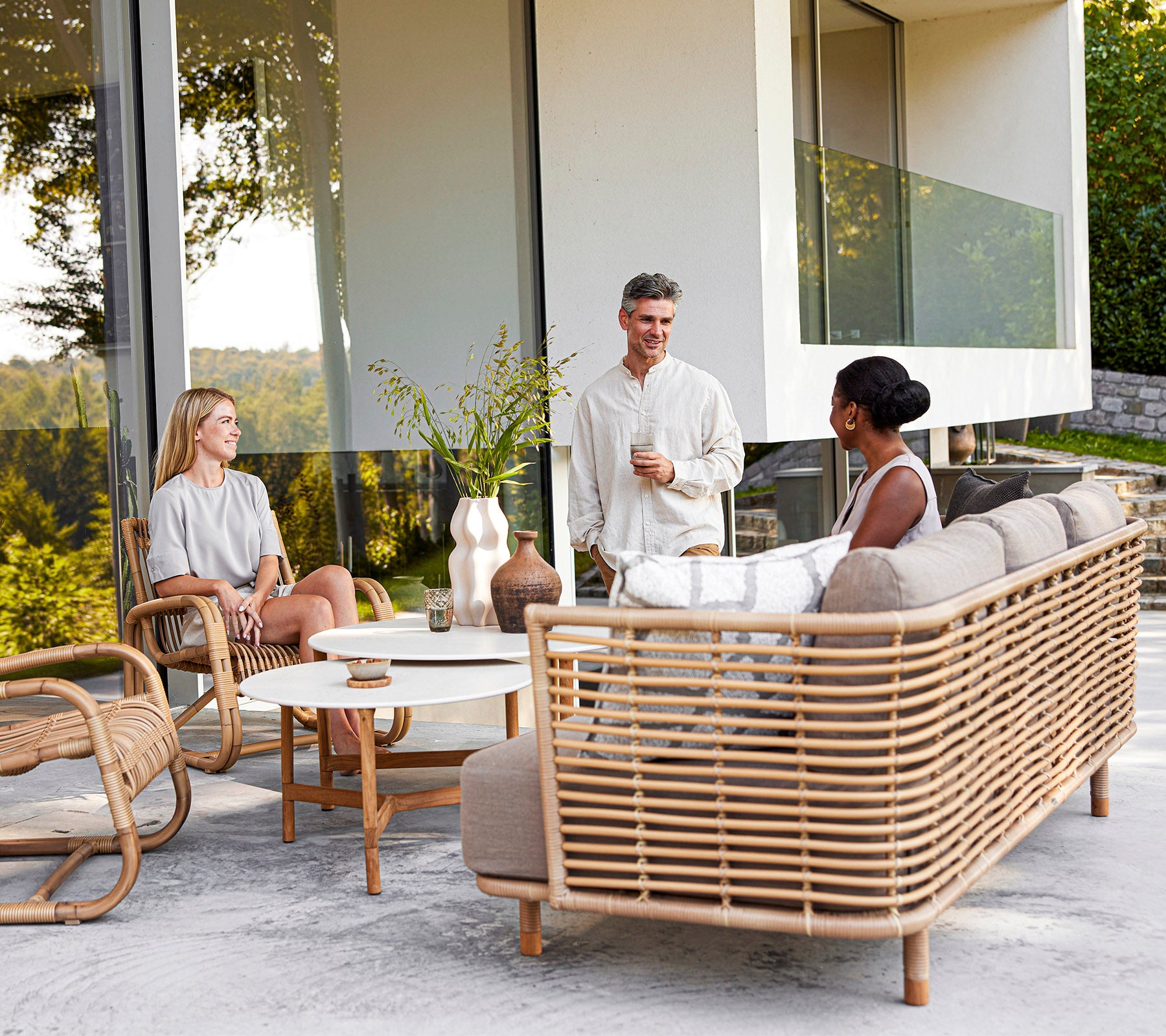 Three people enjoy a casual conversation on stylish rattan furniture with decorative elements, surrounded by greenery. 