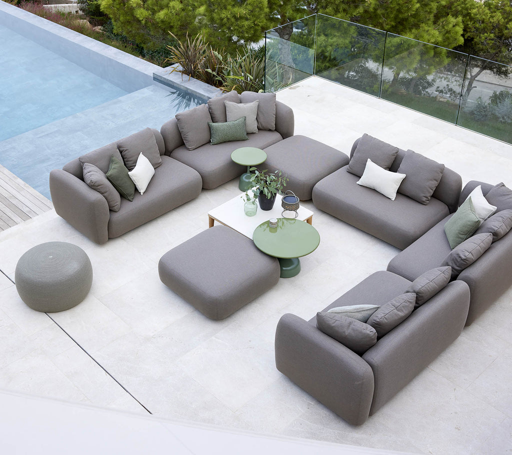 Modern outdoor seating arrangement with gray sofas and green tables beside a pool, surrounded by greenery.