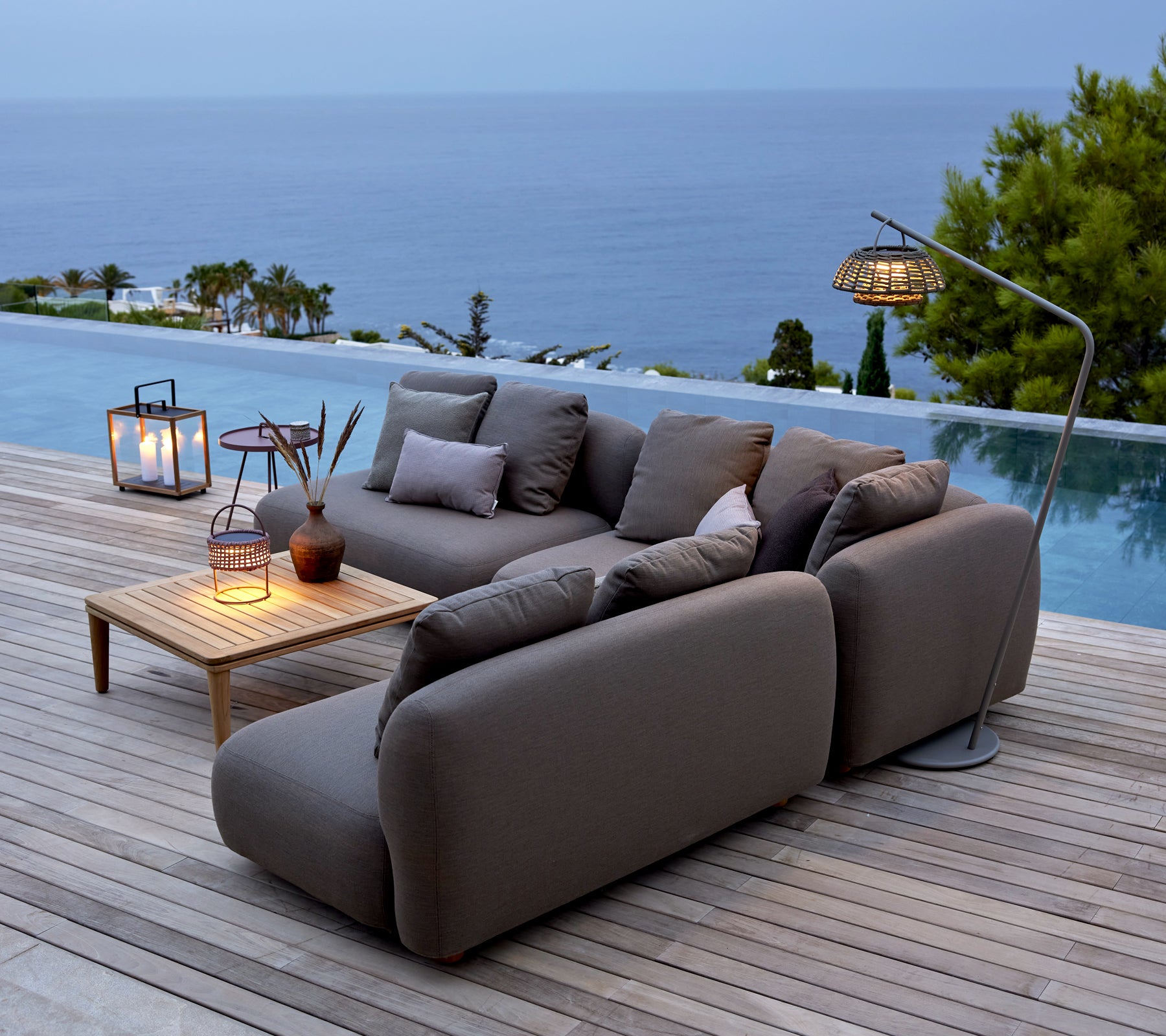 Grey sofa with wooden table and lanterns by a poolside overlooking the ocean.