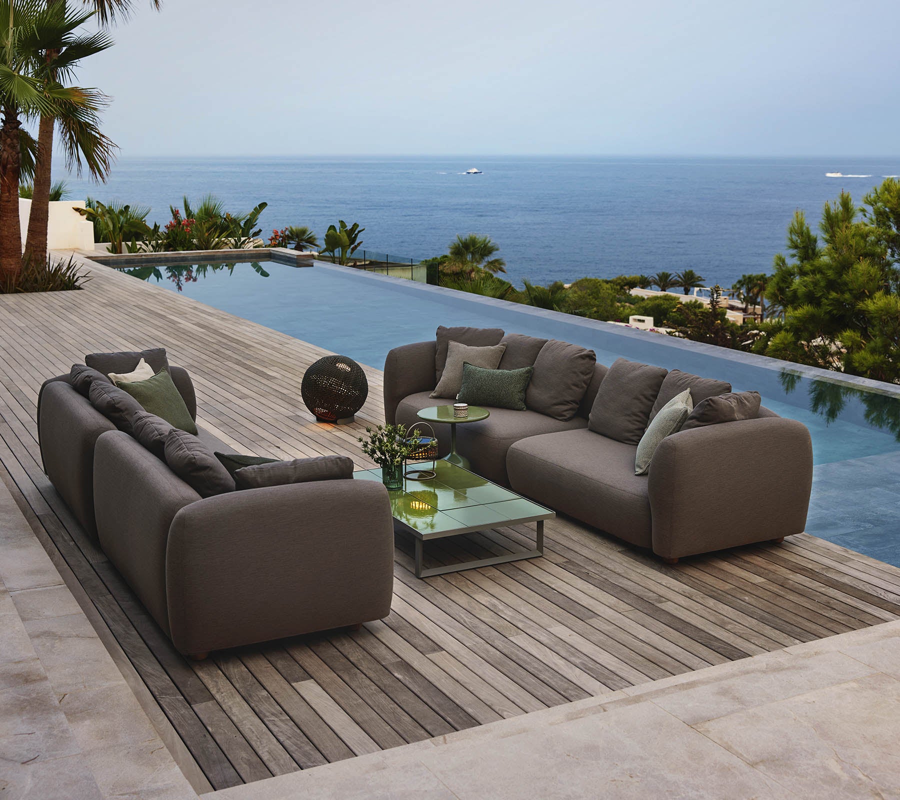 Modern lounge seating arranged around a glass coffee table, overlooking a serene pool and expansive ocean view, surrounded by greenery.