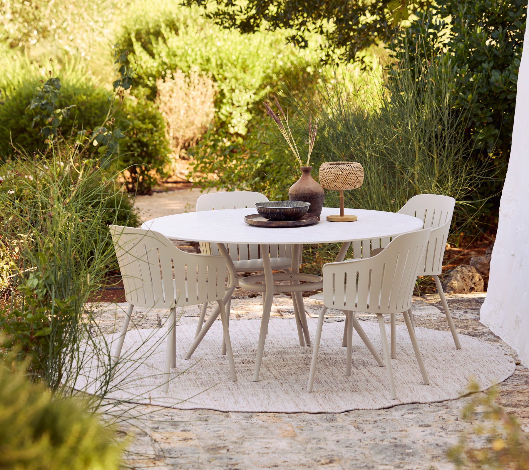 Outdoor dining set with a white round table and beige chairs surrounded by greenery.
