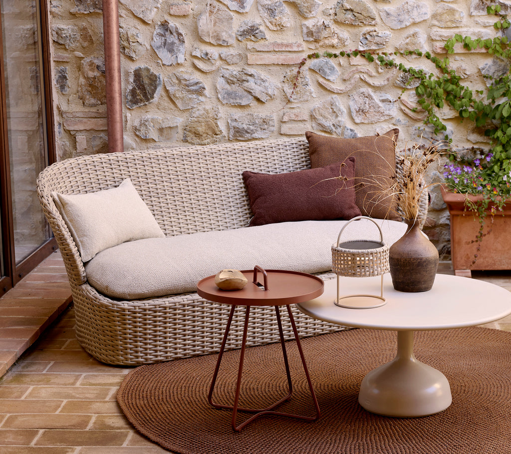 A modern two-person sofa with decorative pillows, paired with a circular coffee table and plants, set against a stone backdrop.