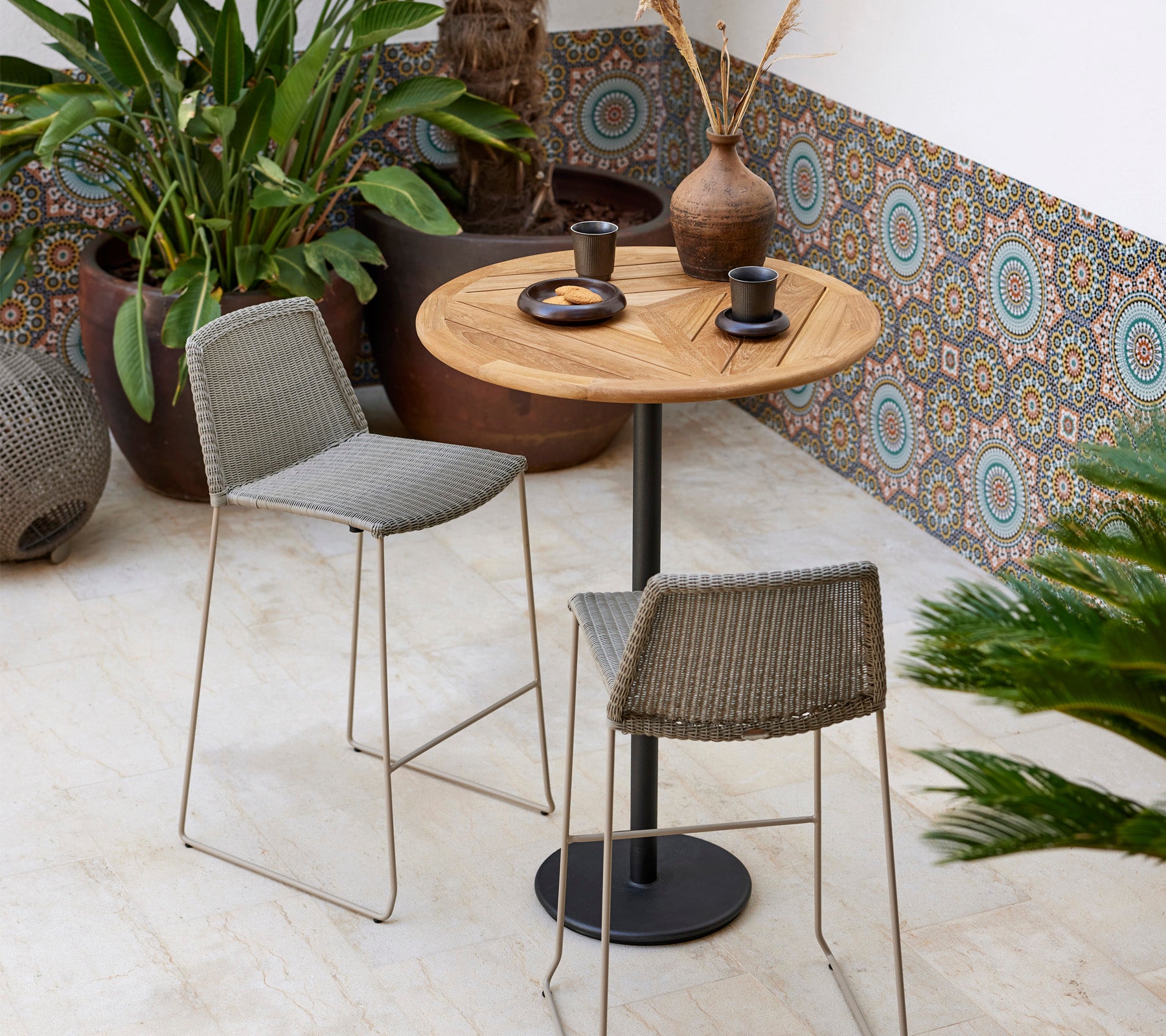 A stylish bistro set featuring a round wooden table and two woven stools, accompanied by decorative plants and modern tableware.