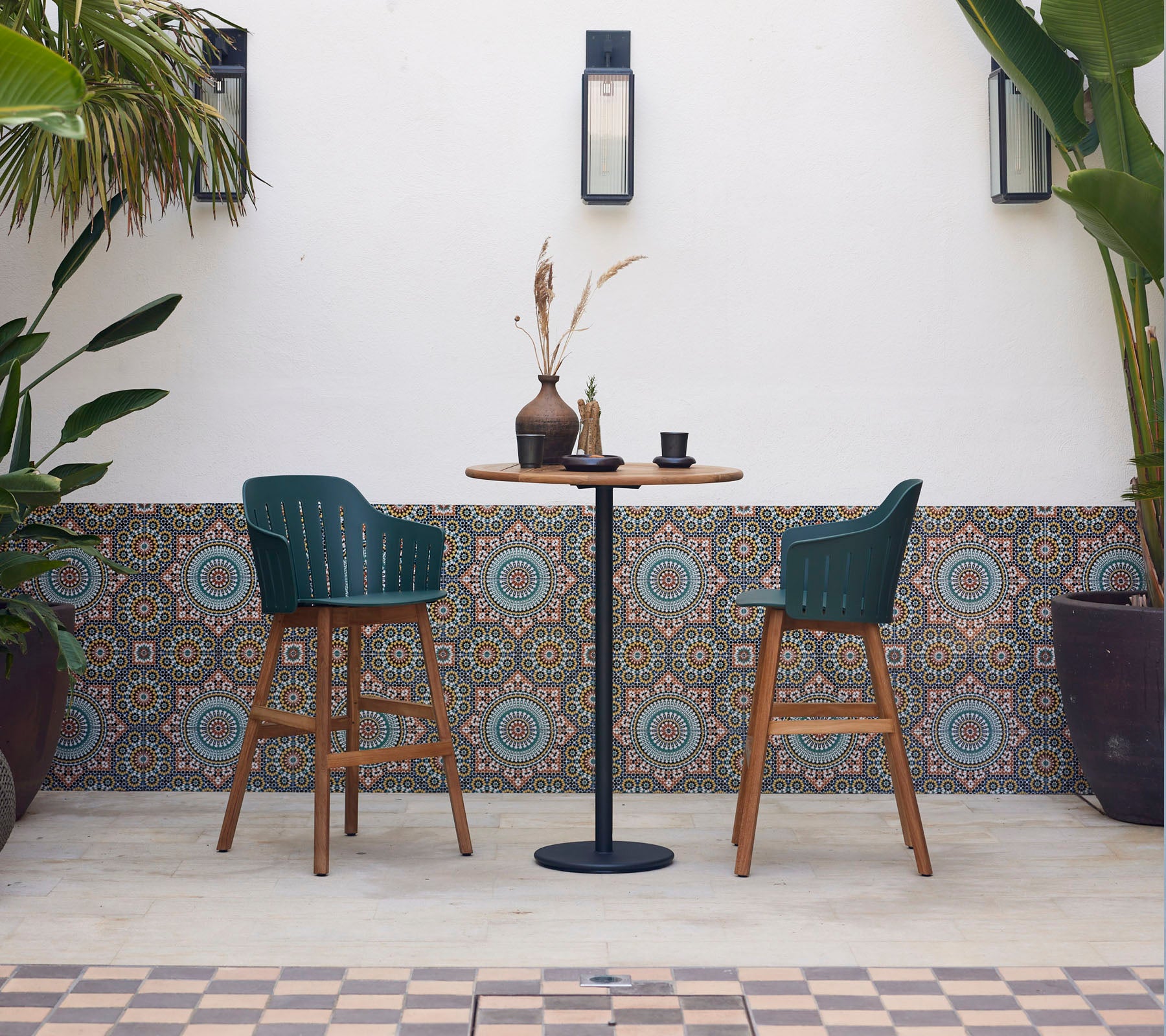 A stylish bar table with two green chairs, decorated with a vase and cup, surrounded by patterned tiles and lush plants.