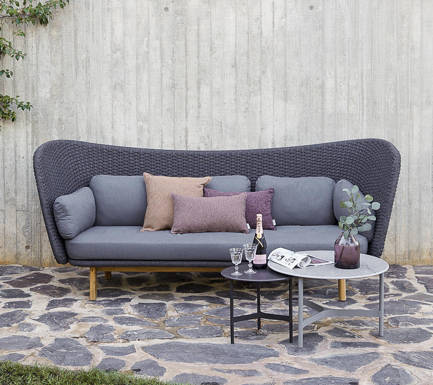 A modern sofa in grey with decorative pillows, accompanied by a small table with drinks and a vase, set against a textured wall.