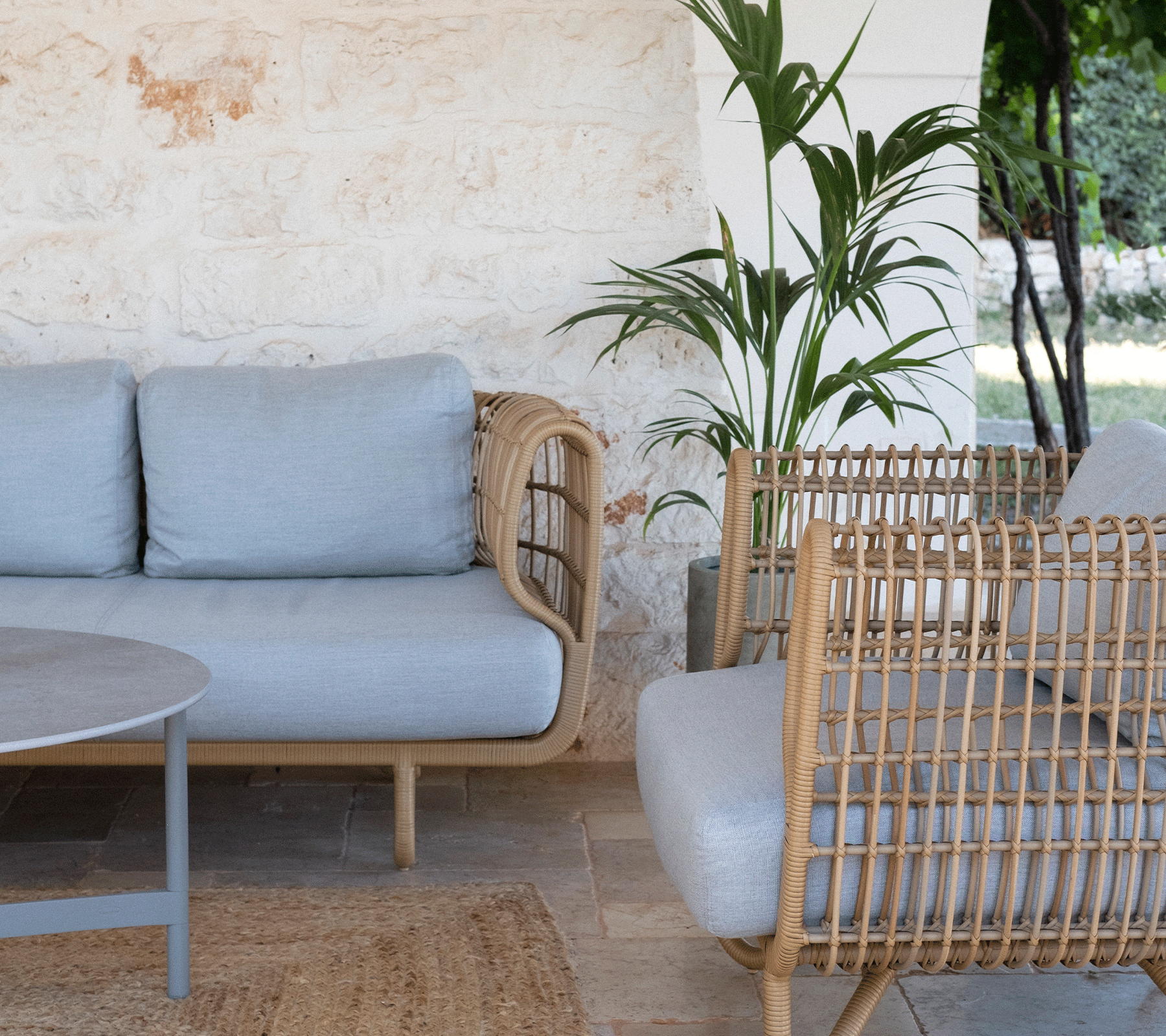 Cozy seating area featuring a light grey sofa, woven armchairs, a round table, and greenery in the background. Suitable for relaxation.