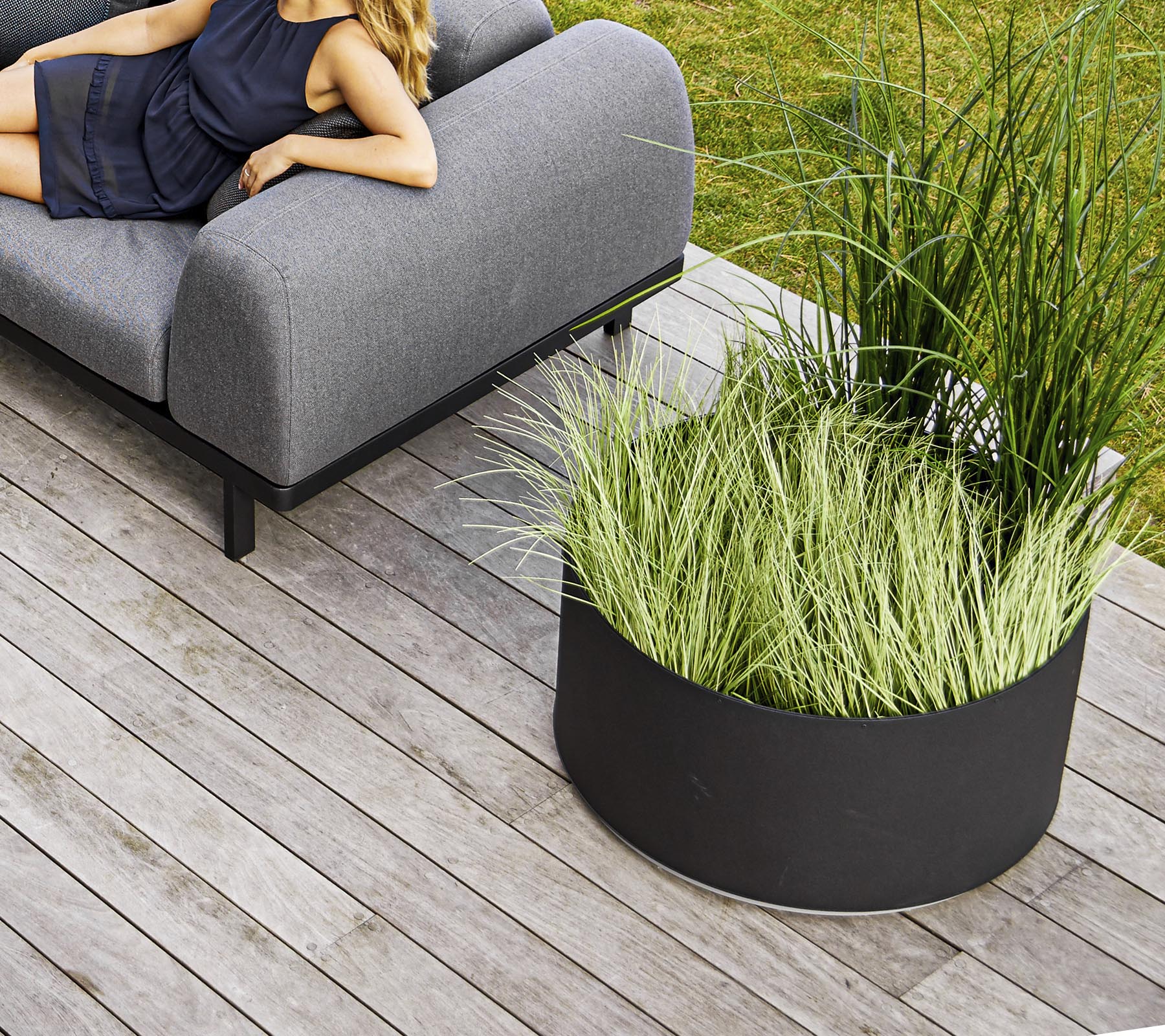A stylish grey sofa beside a circular black planter filled with tall green grass, set on wooden decking.