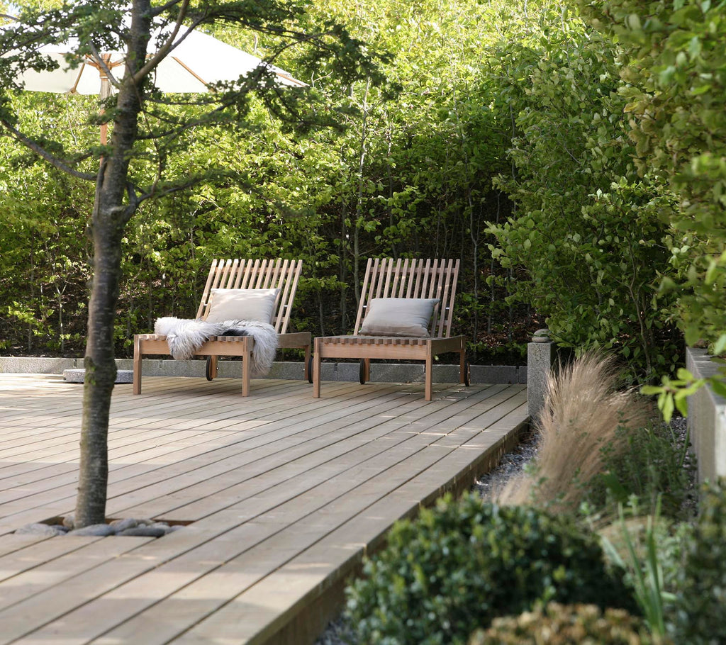 Two wooden lounge chairs with cushions are set on a wooden deck, surrounded by lush greenery and decorative plants.