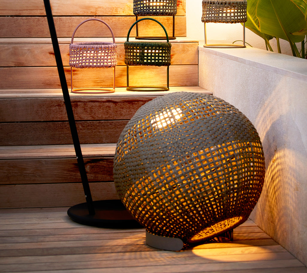 Round taupe illusion lamp with a woven design, casting a warm glow beside stylish lanterns on a staircase.