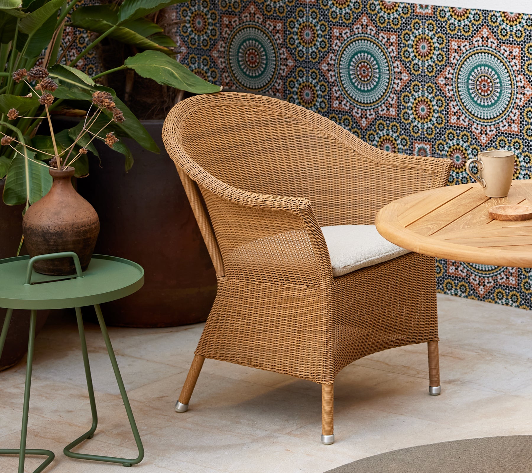 A wicker chair with a cushion beside a green side table, featuring a decorative pattern in the background and a coffee cup on the table.