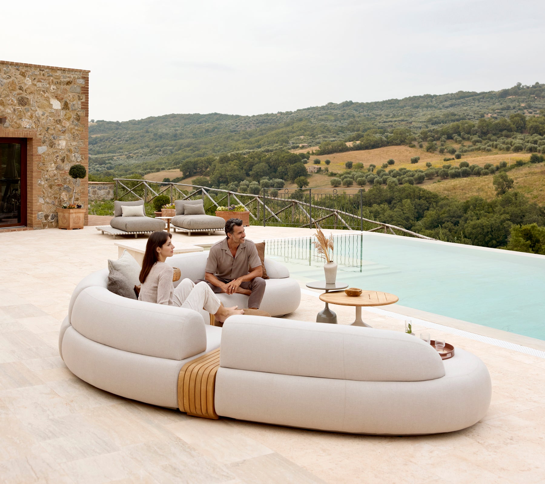 A couple relaxes on a curved, modern sofa beside a stylish coffee table, with a scenic view of rolling hills in the background.