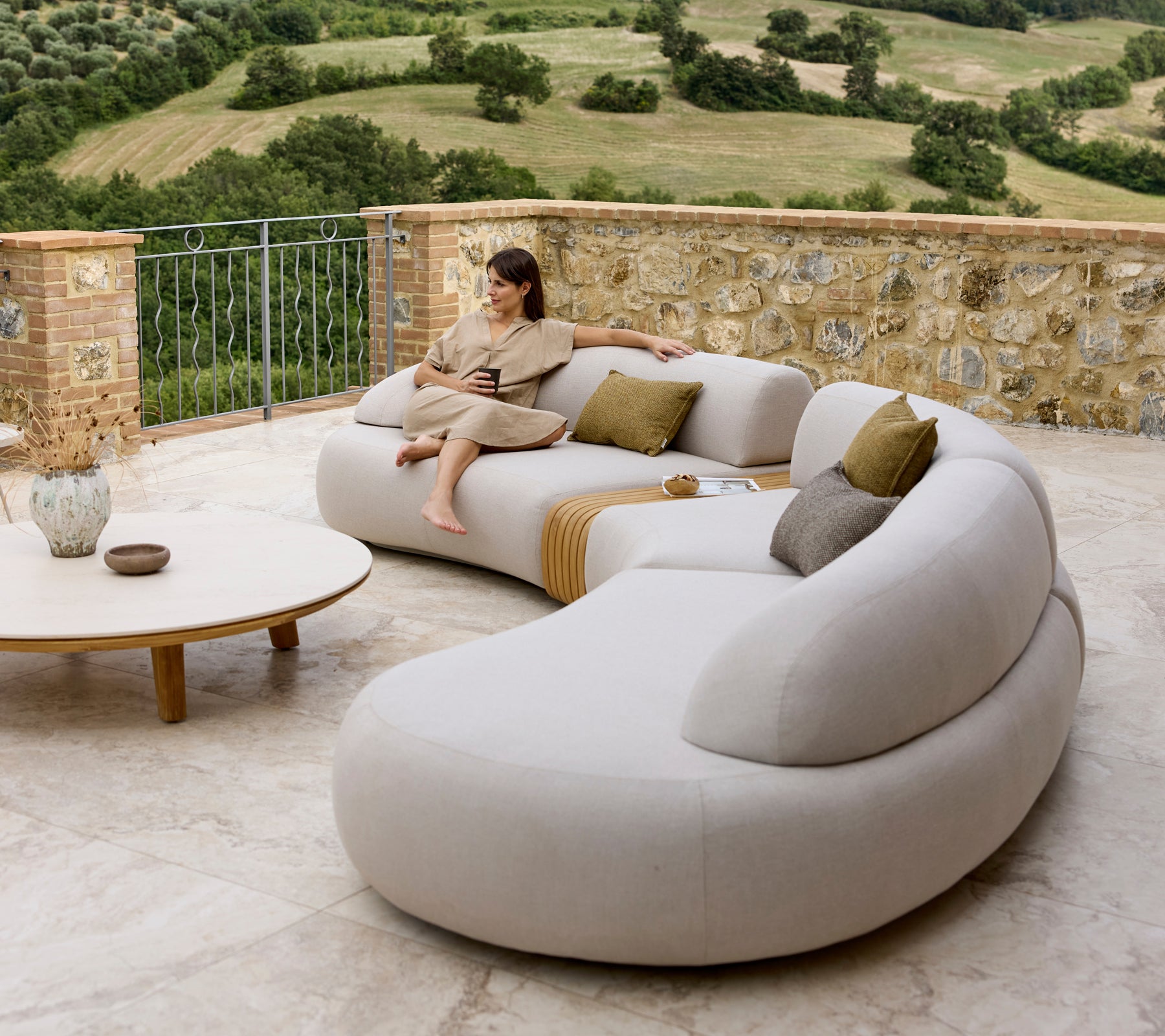 A stylish lounge sofa with soft curves and pillows, complemented by a round coffee table, set against a scenic view.