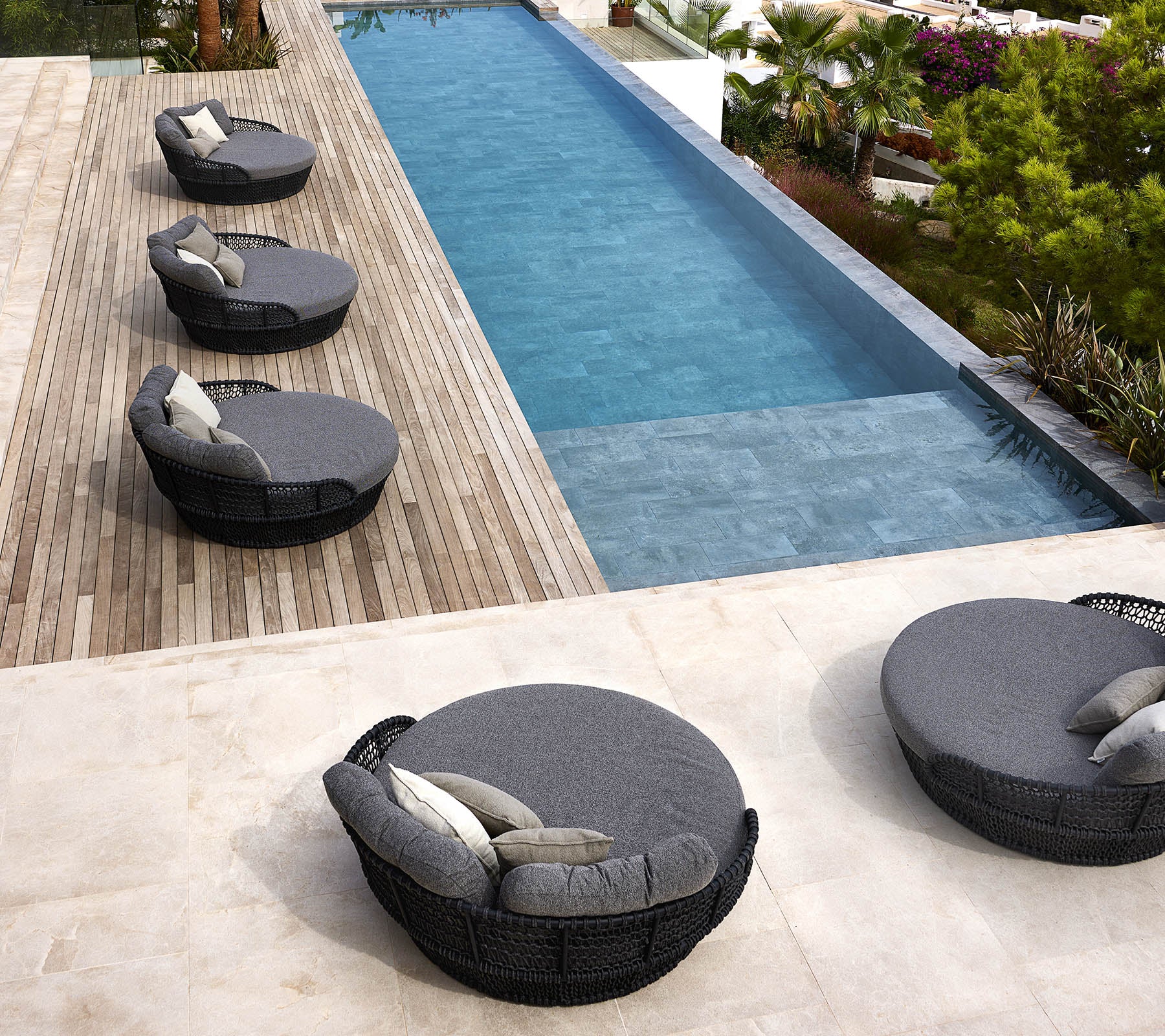 Four round ocean daybeds with cushions are arranged beside a sleek swimming pool surrounded by greenery and wooden decking.