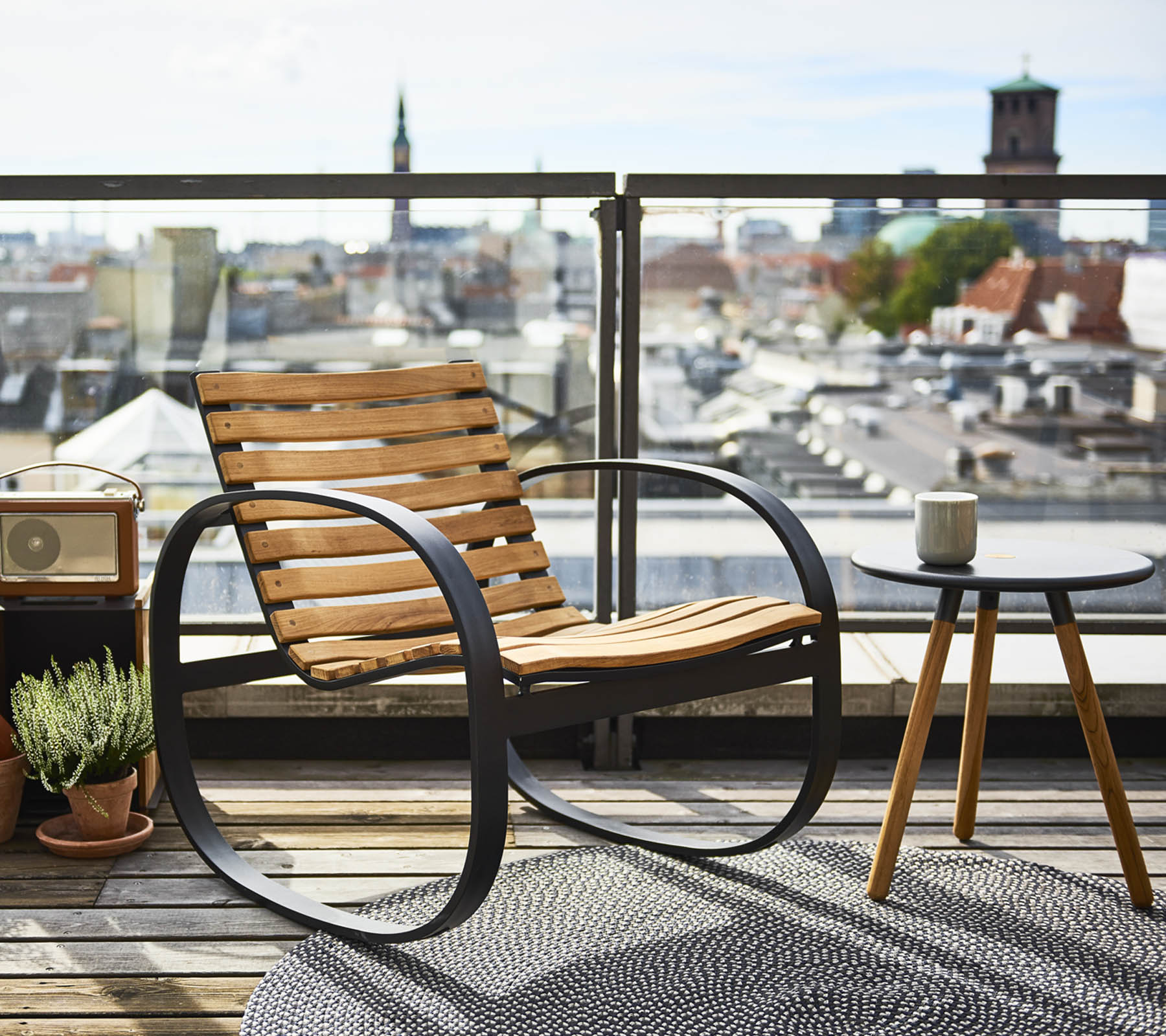 A stylish rocking chair next to a small side table, set against a panoramic city view, adorned with a plant and a rug.