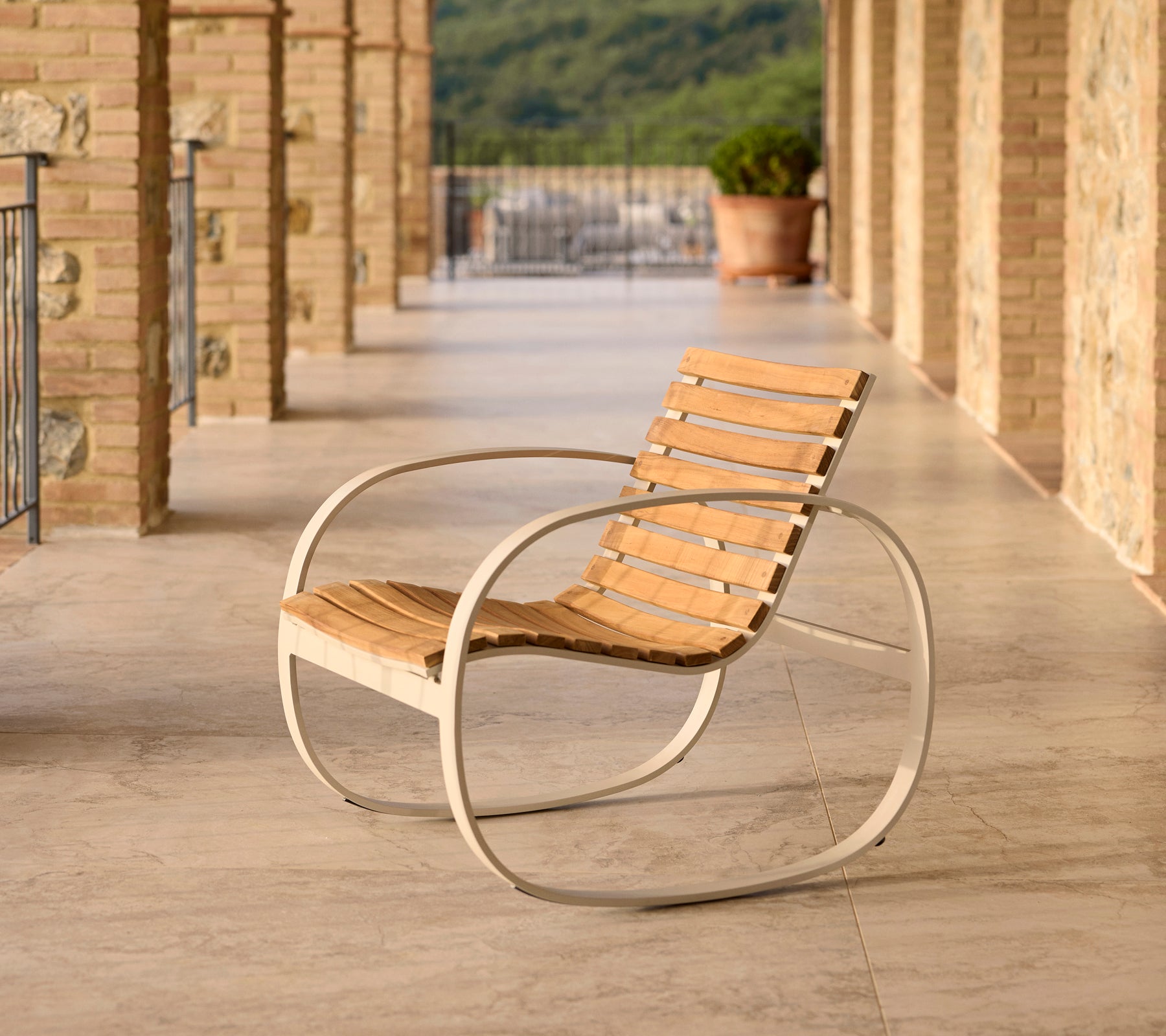 Parc rocking chair with an aluminum frame and wooden slats, positioned on a patio with stone flooring and a scenic backdrop.