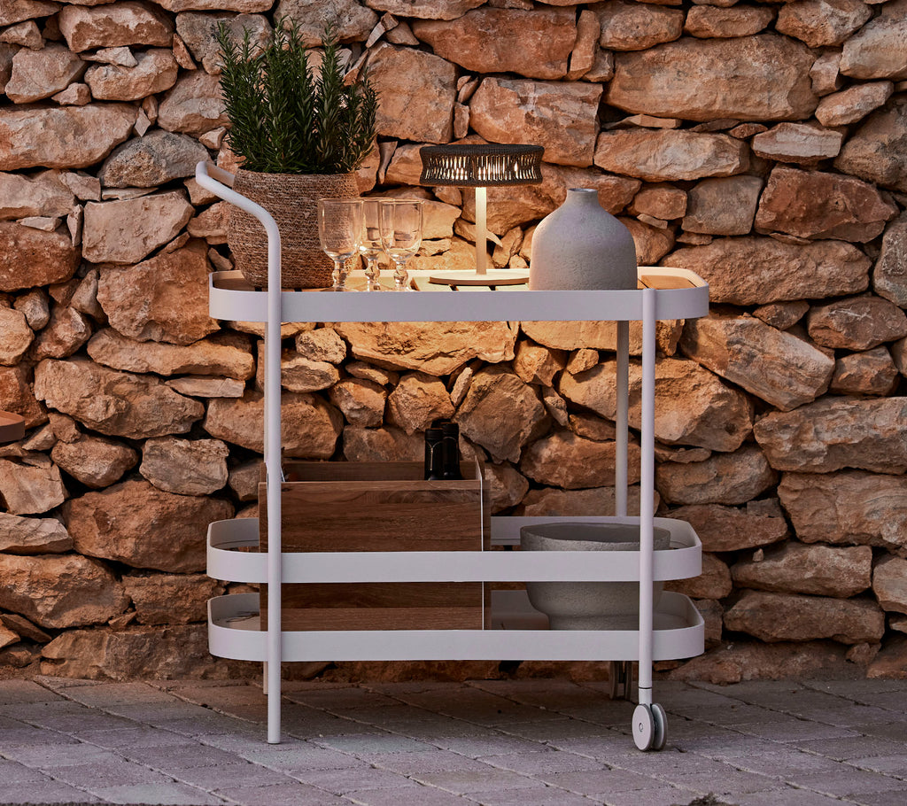 A stylish cart featuring a wooden top, white frame, decorative pot, and sleek lighting against a stone wall backdrop.